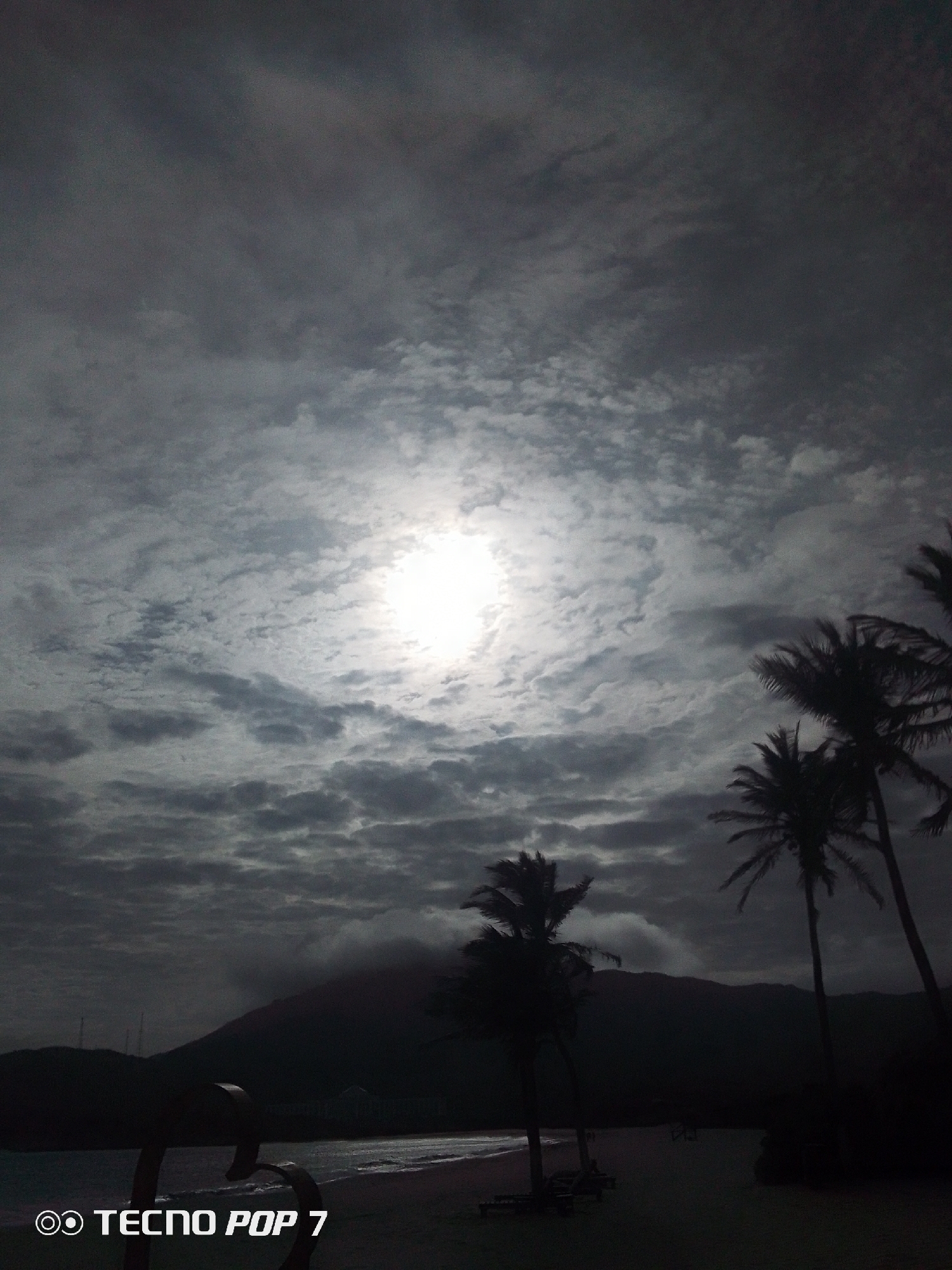 A beach scene in Pedro González, Venezuela, under an overcast sky with the sun weakly visible through the clouds. Palm trees are silhouetted against the sky on the right side of the frame. A dark mountain range is visible in the background. The beach and foreground are dark. "TECNO POP 7" is visible at the bottom of the image.