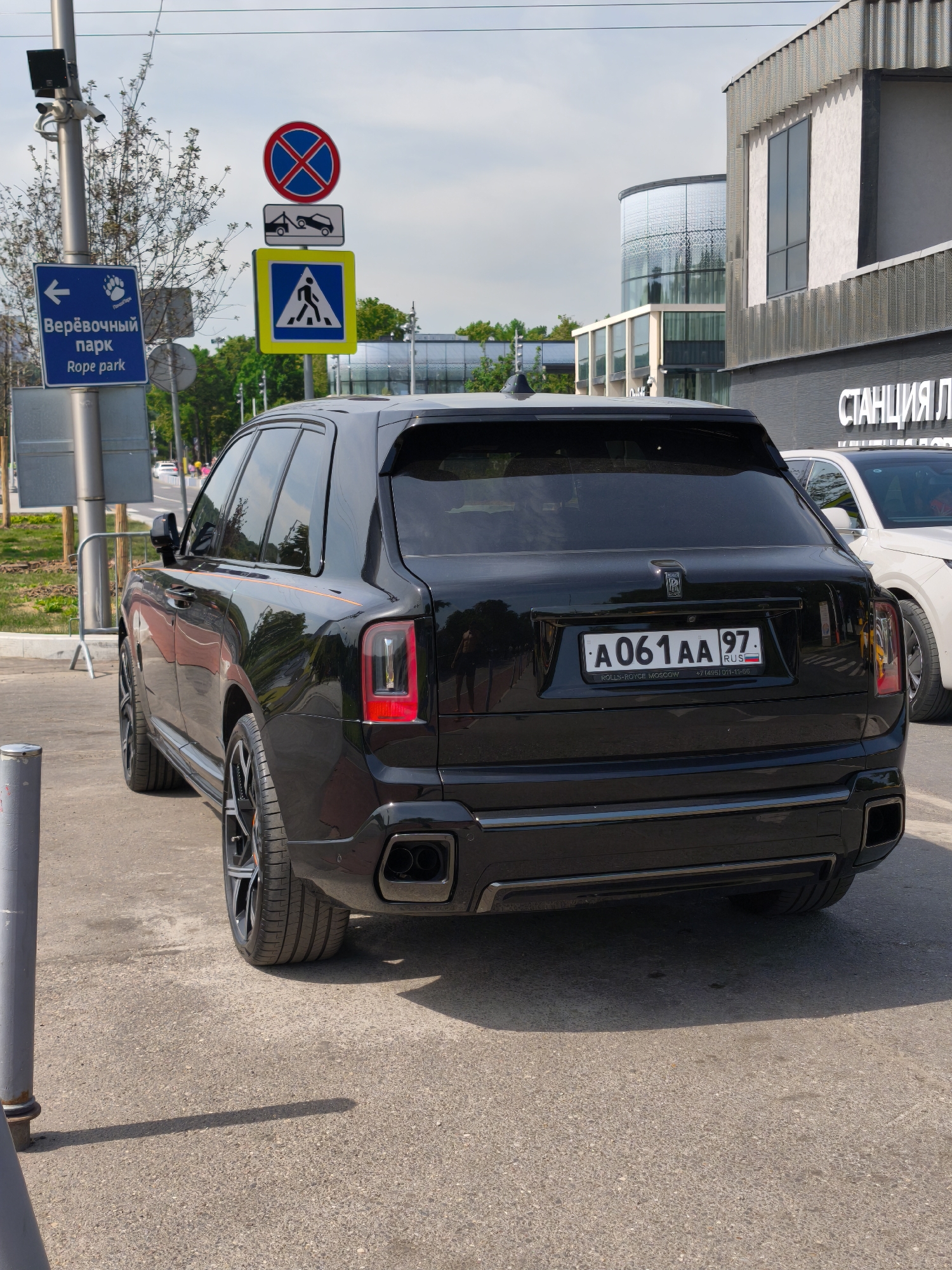 A black Rolls-Royce Cullinan luxury SUV, bearing Russian license plate A061AA 97 RUS (indicating Moscow region 97), is parked facing away from the viewer on an asphalt surface in Moscow, Russia. The vehicle features multi-spoke black wheels and integrated dual rectangular exhaust tips. A subtle orange pinstripe runs along its side body.
To the left, a tall metal pole displays multiple signs. A blue rectangular sign with a white arrow and a bear paw icon reads "Веревочный парк" (Rope park) and "Rope park" below it, indicating a recreational area. Stacked above it are three road signs: a red circle with a blue 'X' (No Stopping), a white rectangular sign depicting a tow truck (No Stopping, Tow-Away Zone), and a blue square with a white triangle containing a walking person (Pedestrian Crossing). Security cameras are mounted on the pole.
In the background, modern architectural buildings are visible under an overcast sky. One building features a curved, reflective glass facade, while another has a textured grey and white exterior with large vertical windows. A partial sign "СТАНЦИЯ Л" (STANTSIYA L...) is visible on the building to the right. A white SUV is partially parked behind the Rolls-Royce. Reflections of individuals are discernible in the darkened windows of the black SUV. The scene is set in a developed urban environment, likely near a public park or recreational complex given the "Rope Park" signage.