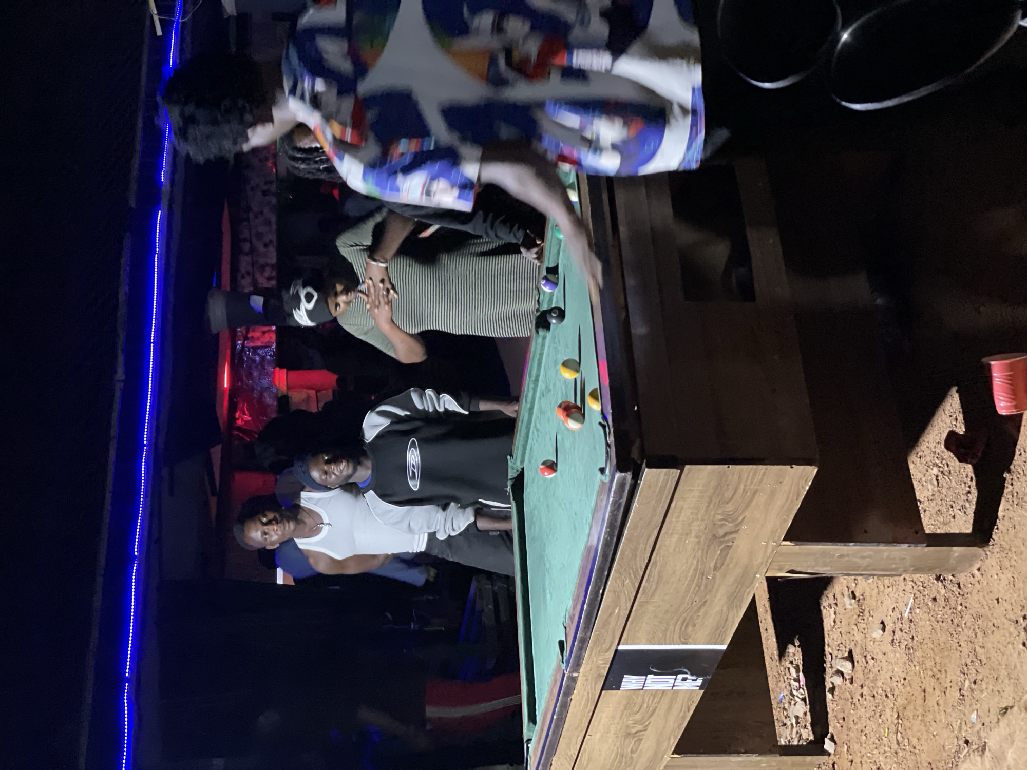 The image depicts a nighttime scene at an outdoor establishment with a pool table in Durumi, Nigeria. Four individuals are visible around the pool table. Two men stand behind the table, one in a white tank top and another in a black and grey long-sleeved shirt. A woman in a striped green dress is positioned behind the table, facing towards it. Another person, partially obscured, is leaning over the pool table, seemingly playing. The environment appears to be somewhat rustic, with a dirt or gravel ground surrounding the table and a covered structure with blue and red lighting overhead. There are several pool balls on the table, and some appear to be in play.