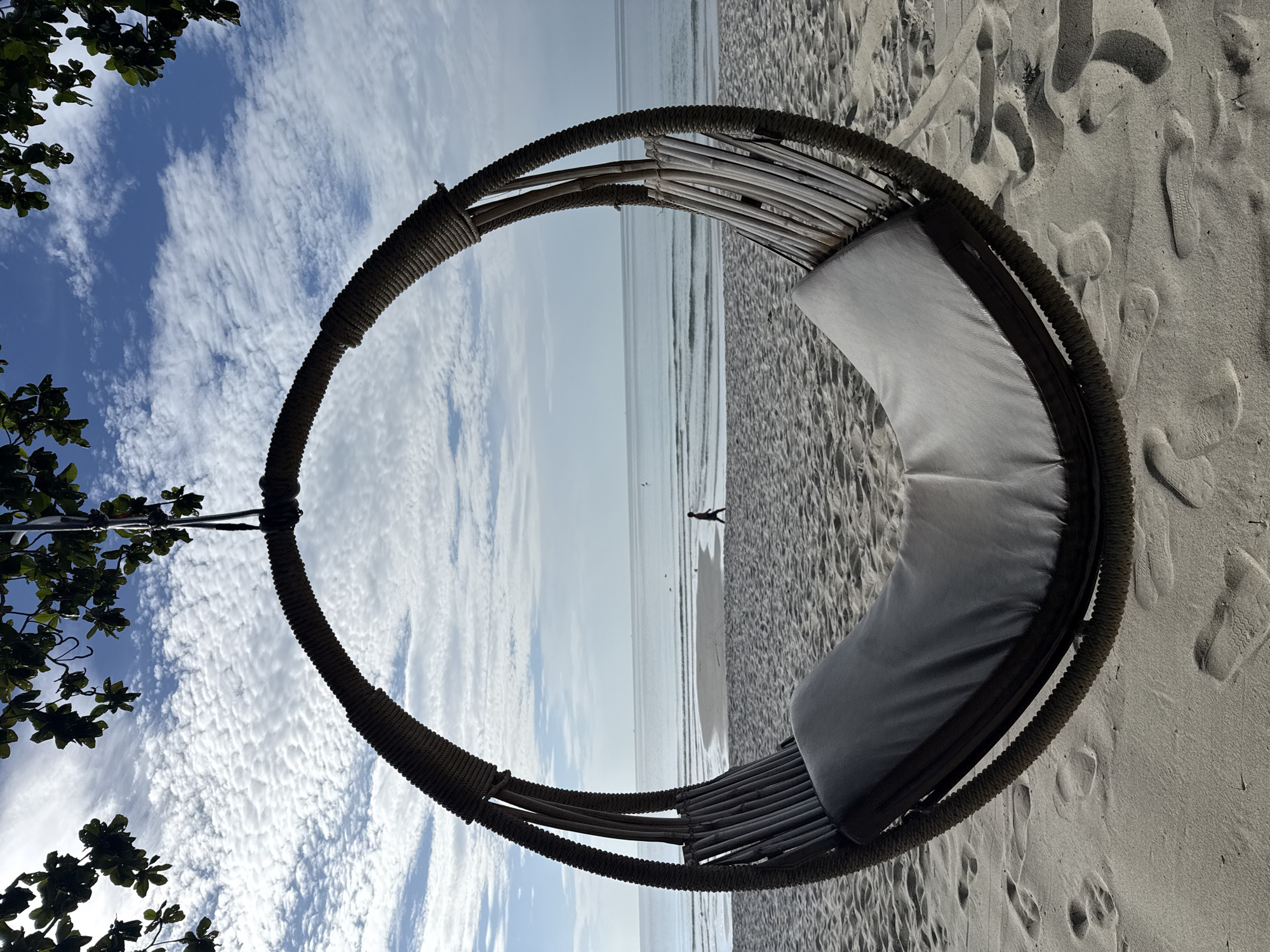 A circular rope swing with a grey cushion is suspended in the foreground, positioned on a sandy beach. The swing faces a calm ocean with gentle waves and a light blue sky filled with scattered white clouds. In the background, a lone figure walks along the shoreline. Trees with green leaves frame the top of the image. The scene appears to be on a beach in Ko Samui District, Thailand.