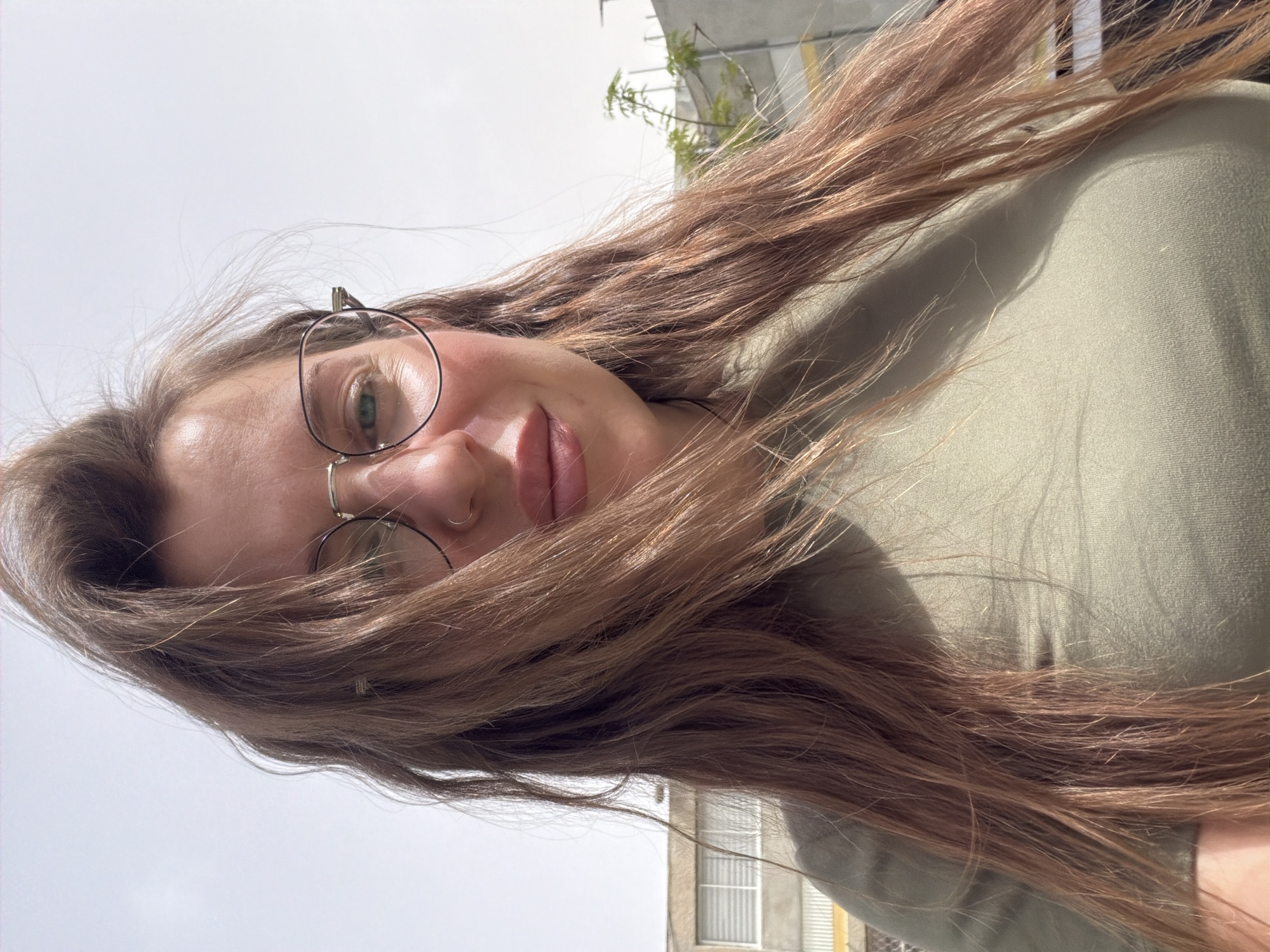 A woman with long, light brown hair and wearing round glasses is facing the camera. She is wearing an olive green top. She appears to be outdoors during the day, with a building visible in the background. The location appears to be Bat Yam, Israel. The camera angle is low.