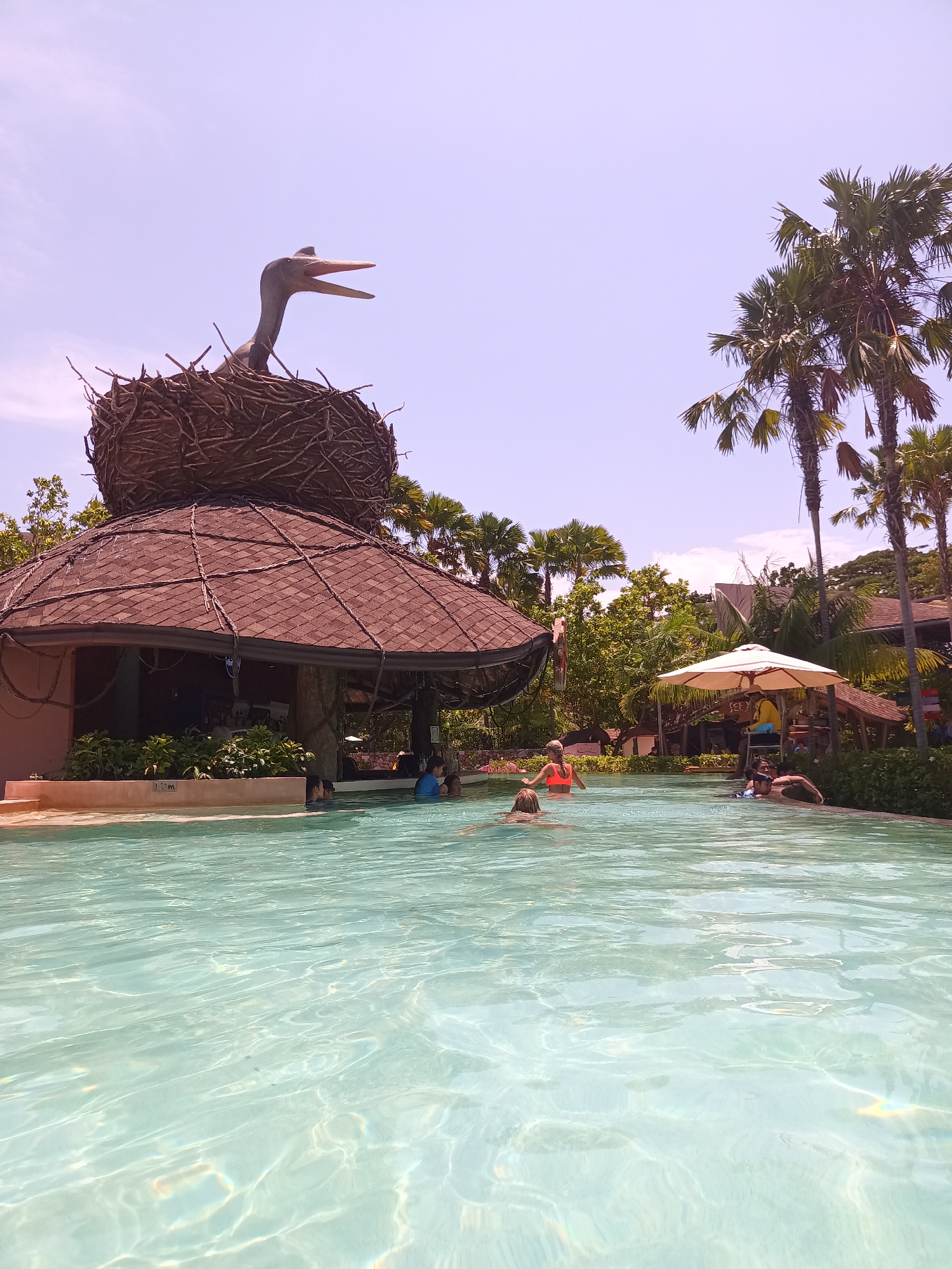 The image depicts a water park scene in Hua Hin City Municipality, Thailand, on a sunny day. The clear, light blue water of a pool takes up a significant portion of the foreground, with visible ripples and light reflecting off the surface. In the middle ground, several people are swimming, including some wearing orange life jackets. To the left, a structure resembling a thatched-roof hut houses a bar area. Atop this hut sits a large nest with a sculpted bird, possibly a heron or stork, with its beak open. Lush tropical foliage, including palm trees, frames the pool area and the structure. A white sun umbrella provides shade to a lifeguard in a tall chair. The sky above is a light blue with a slight purple hue.