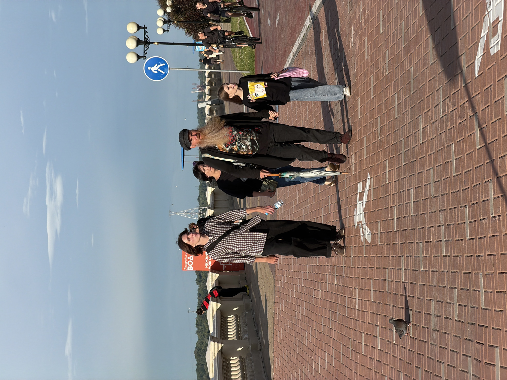 This image captures a sunny day on a brick-paved promenade in Nizhny Novgorod, Russia. In the foreground, a young person with long brown hair walks alone, wearing a plaid shirt and dark pants, holding a water bottle. Further back, a family strolls together: a woman with dark hair carries an umbrella, a man with a long beard and a cap wears a graphic t-shirt and dark clothing, and a young girl in a black sweatshirt and jeans holds his hand and a pink bag. In the background, a man in a red and black striped shirt leans over a decorative stone wall. Cyclists are visible in the distance on a separate path, and streetlights and a pedestrian crossing sign are present. The sky is partly cloudy with a bright blue hue.