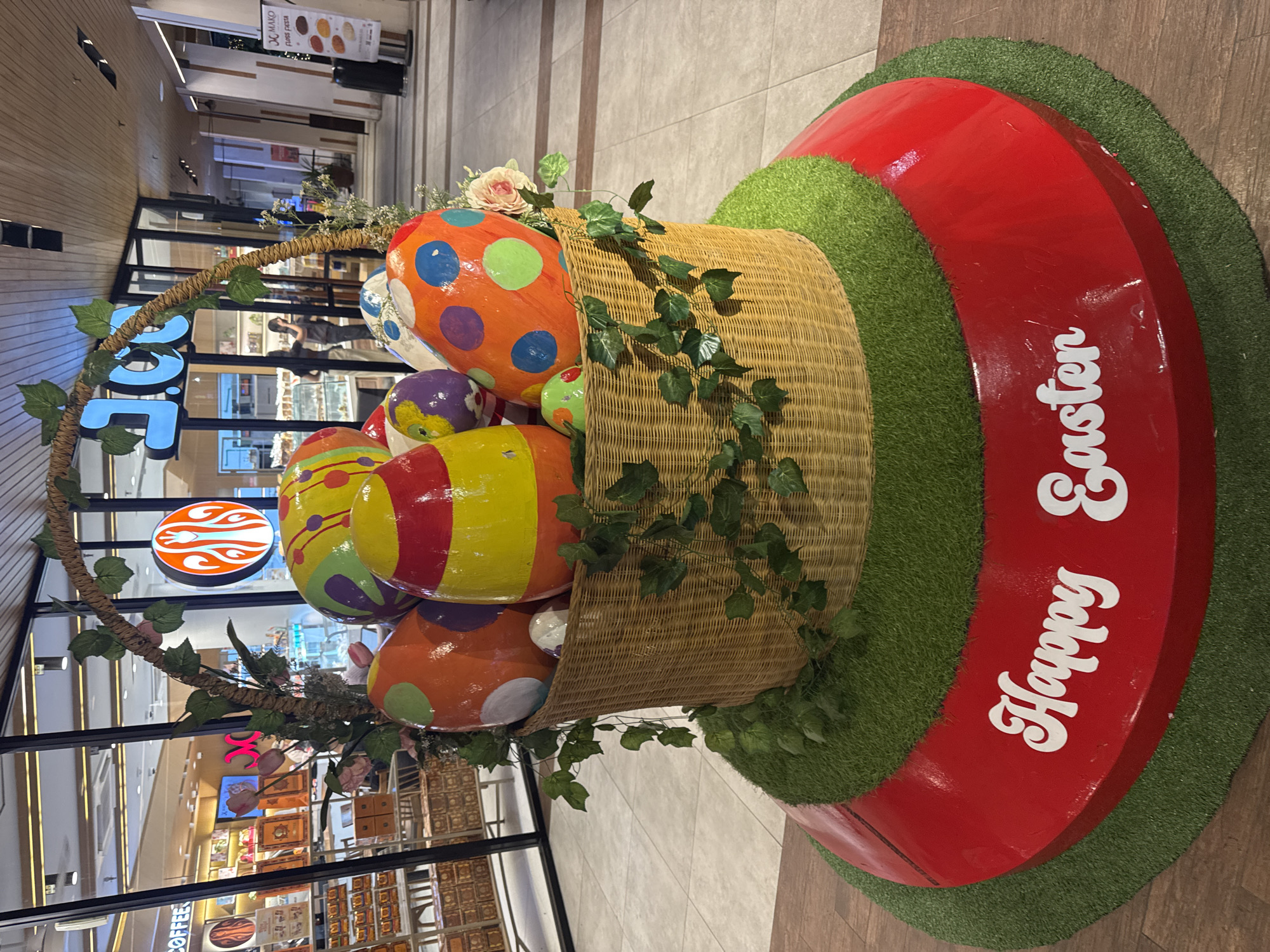 A large Easter display stands in a mall in Kuta Selatan, Indonesia. The display consists of a woven basket overflowing with brightly colored, oversized Easter eggs, decorated with polka dots, stripes, and other patterns. The basket has a handle made of rope covered in leaves, and is surrounded by greenery. The display is set on a circular base of red and green, with "Happy Easter" written on the red portion in white lettering. The surrounding mall area includes storefronts with glass windows, one with "COFFEE" visible and another with a round orange and white logo. Shoppers and customers can be seen inside the storefronts. The mall floor is tiled in a light brown pattern.