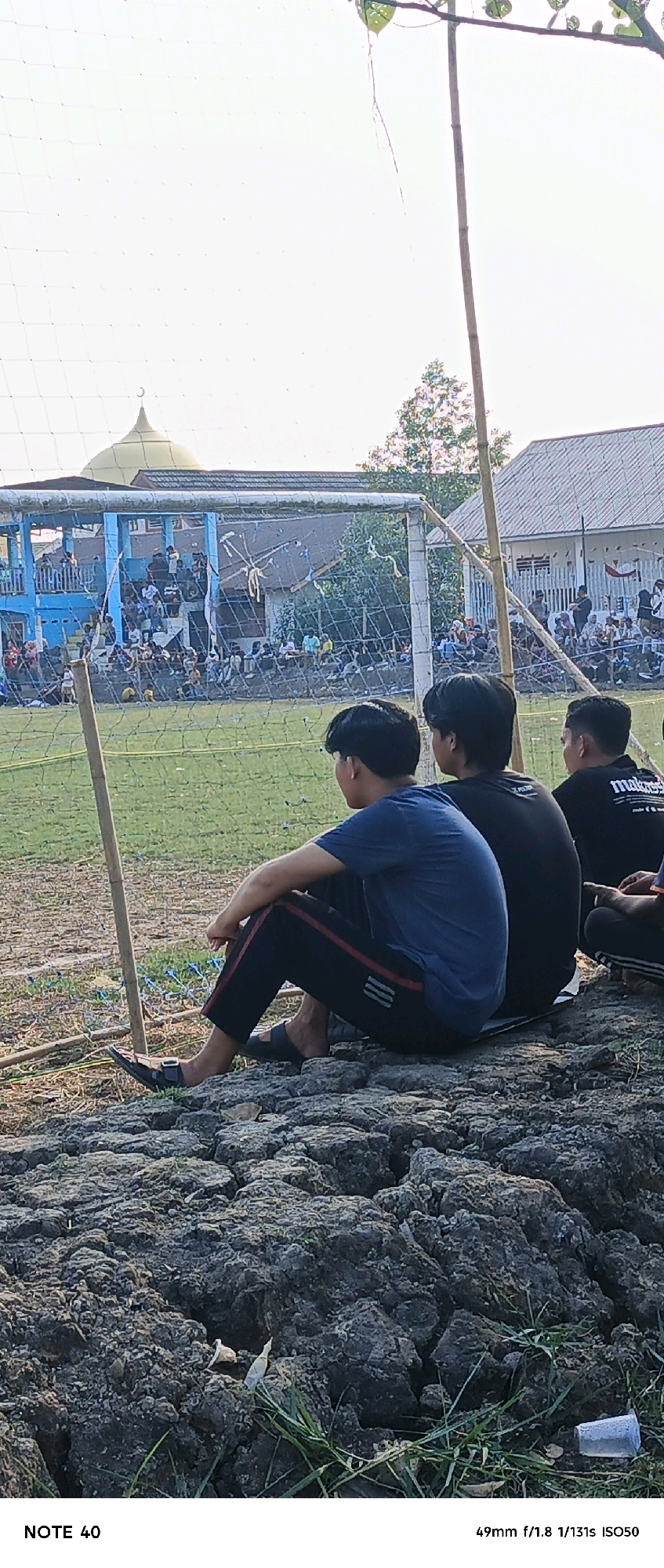 The image depicts a soccer field in Simbur Naik, Indonesia, with a crowd of spectators in the background, seated in bleachers. A mosque with a dome is visible behind the bleachers. In the foreground, three young men are seated on the ground, facing away from the camera, watching the game. The ground in the foreground is rocky and dry, with sparse grass. The overall lighting suggests it is daytime, likely late afternoon.