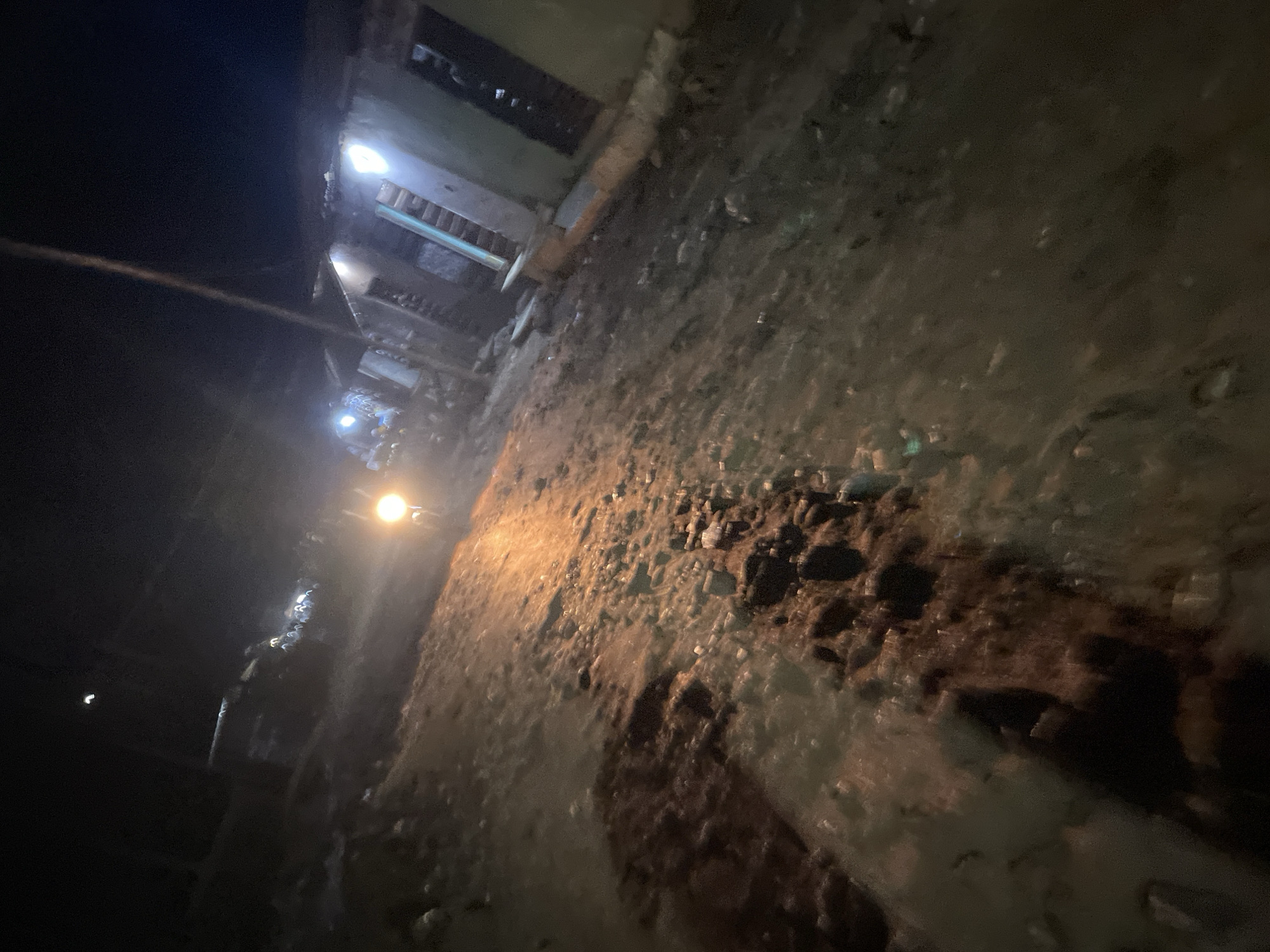 The image depicts a nighttime scene on a dirt road in Lokoja, Nigeria. The road is muddy and uneven, with visible debris and puddles of water reflecting the light from a vehicle's headlights. The headlights of a motorcycle are illuminated in the center of the frame, casting a strong orange glow on the road ahead. In the background, several structures are visible, appearing to be houses or shops, with some lights illuminated from within. A person or group of people can be seen near the illuminated structures, their forms indistinct in the low light. A tall, slender pole or antenna stands vertically in the background, to the right of the motorcycle's light. The overall impression is of a rural or semi-urban setting at night, with limited artificial lighting.