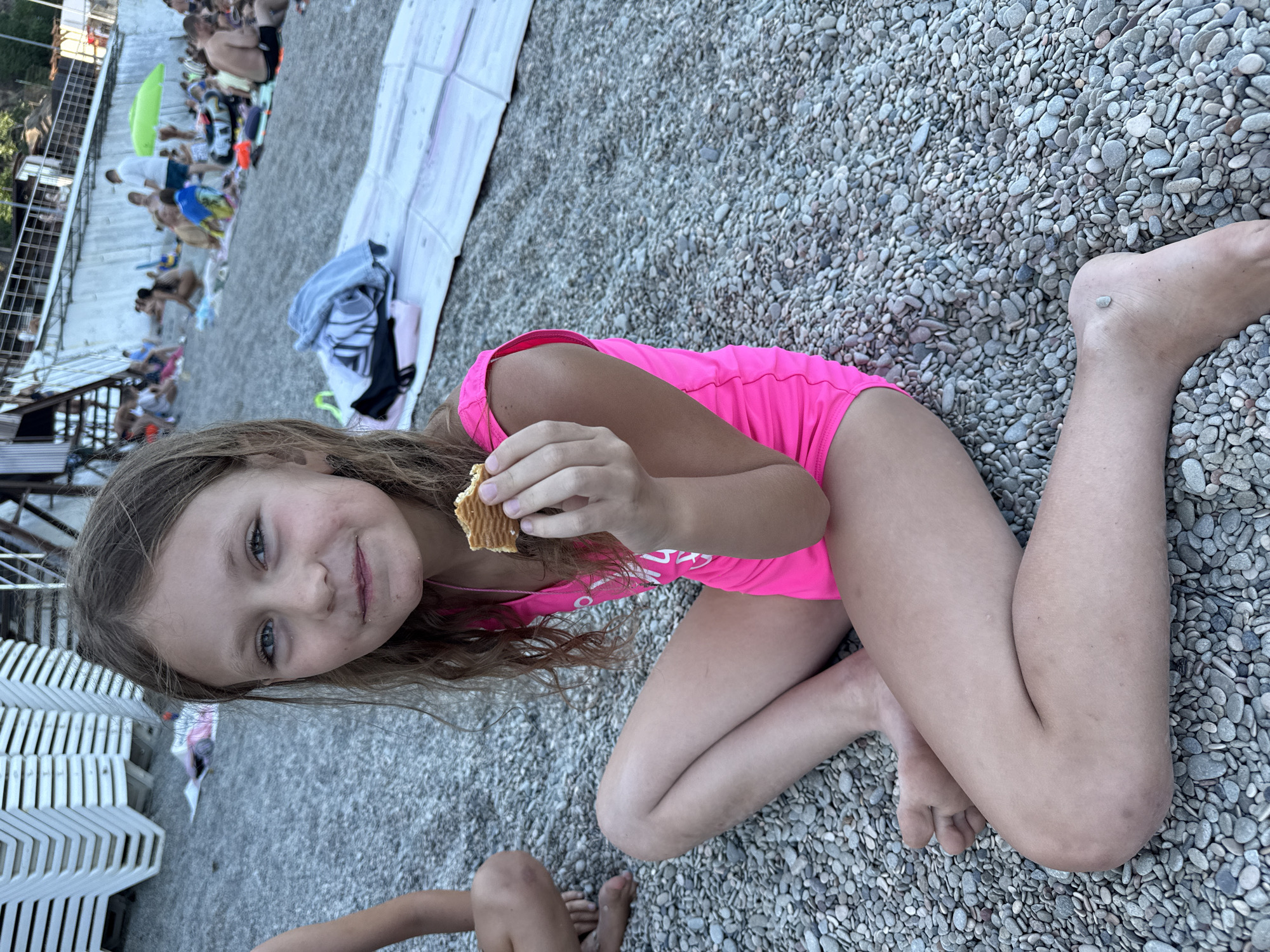 A young girl is lying on a pebble beach in Partenit, Ukraine, wearing a bright pink swimsuit. She is holding a biscuit in her hands and looking at the camera with a slight smile. The girl has long, wavy brown hair and blue eyes. Her legs are bent, and she is resting on her elbows.
In the background, other people can be seen on the beach, some sitting on towels and others standing. There are also stacks of white beach chairs. The overall scene suggests a warm, sunny day at the beach. The lighting indicates it is daytime, likely late afternoon given the soft light. The presence of people suggests a public beach during a recreational period. The environment is characterized by numerous small, grey pebbles covering the ground. The backdrop includes some greenery and structures, possibly part of a beach resort or boardwalk area. No visible text is present in the image.