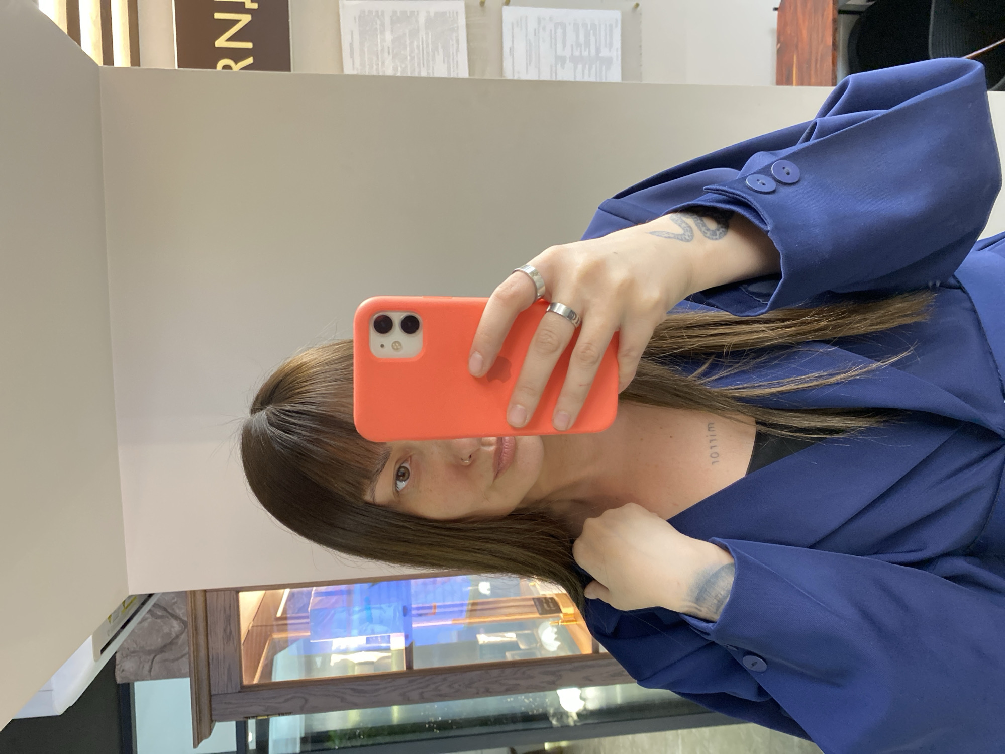 An indoor mirror selfie captures a person, likely a young adult female, with long dark brown hair and bangs, holding an orange smartphone (resembling an iPhone with dual cameras) in her right hand. Her left hand, adorned with two silver rings on the index and middle fingers, also holds the phone. She is wearing a dark blue blazer or jacket with two prominent buttons visible on the cuff of her right sleeve, and a dark top underneath. A small stud nose piercing is visible. Notable tattoos include a snake-like design on her left forearm/wrist and mirrored text or symbols on her right collarbone area.
The background, reflected in the mirror, features a light-colored wall. A partial sign with the letters "RN" is visible at the top. Two white documents or papers are pinned to the wall higher up. Below, a wooden display case with glass showcases reflections of blue and white lights or objects, suggesting a commercial environment such as a store or salon. The overall scene appears to be an interior space in Votkinsk, Russia. The image is oriented 90 degrees clockwise from standard portrait view.