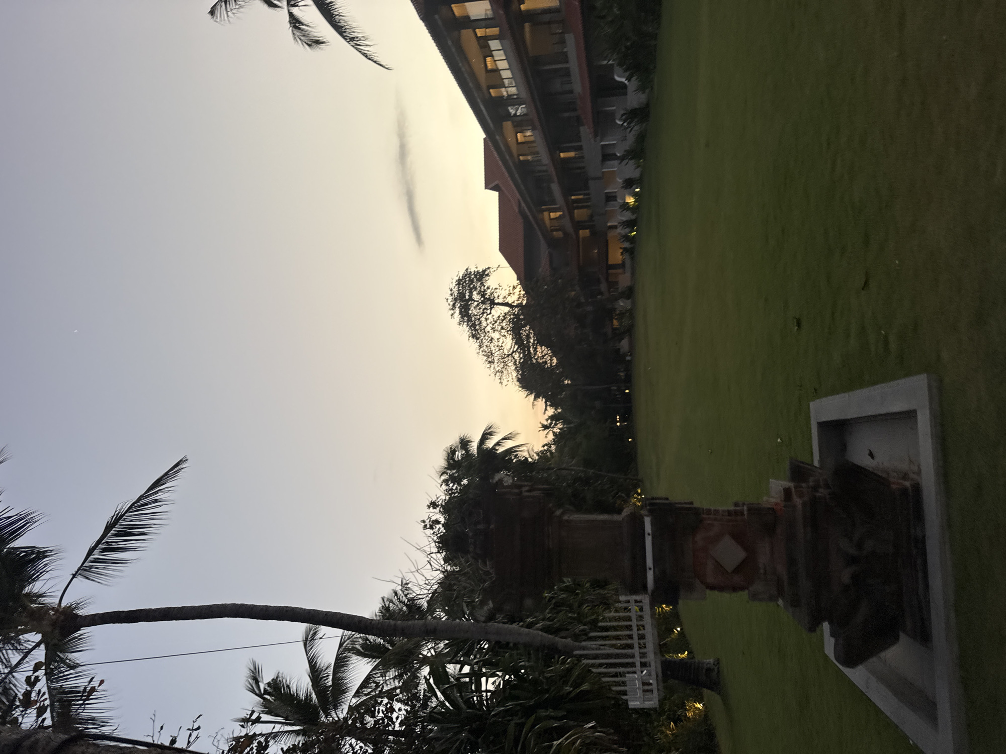 A low-angle, full shot captures a hotel and its manicured lawn at dusk in Kuta Selatan, Indonesia. The sky is a gradient of pale blue at the top, transitioning to a soft orange and yellow glow near the horizon. Silhouettes of palm trees frame the left side of the image, their fronds extending into the sky. In the foreground, a stone pedestal, possibly a decorative monument or an altar, stands on a concrete base, partially obscuring a white fence and a palm tree trunk to its left. Beyond the pedestal, a vast, flat expanse of lush green grass stretches towards a long, multi-story hotel building. The hotel, with its terracotta-tiled roof, features numerous windows and balconies illuminated by interior lights, casting a warm glow. A few scattered trees, including a larger, bare-branched tree to the left of the hotel, add depth to the scene. The overall atmosphere is serene and tropical, characteristic of a resort in southern Bali.