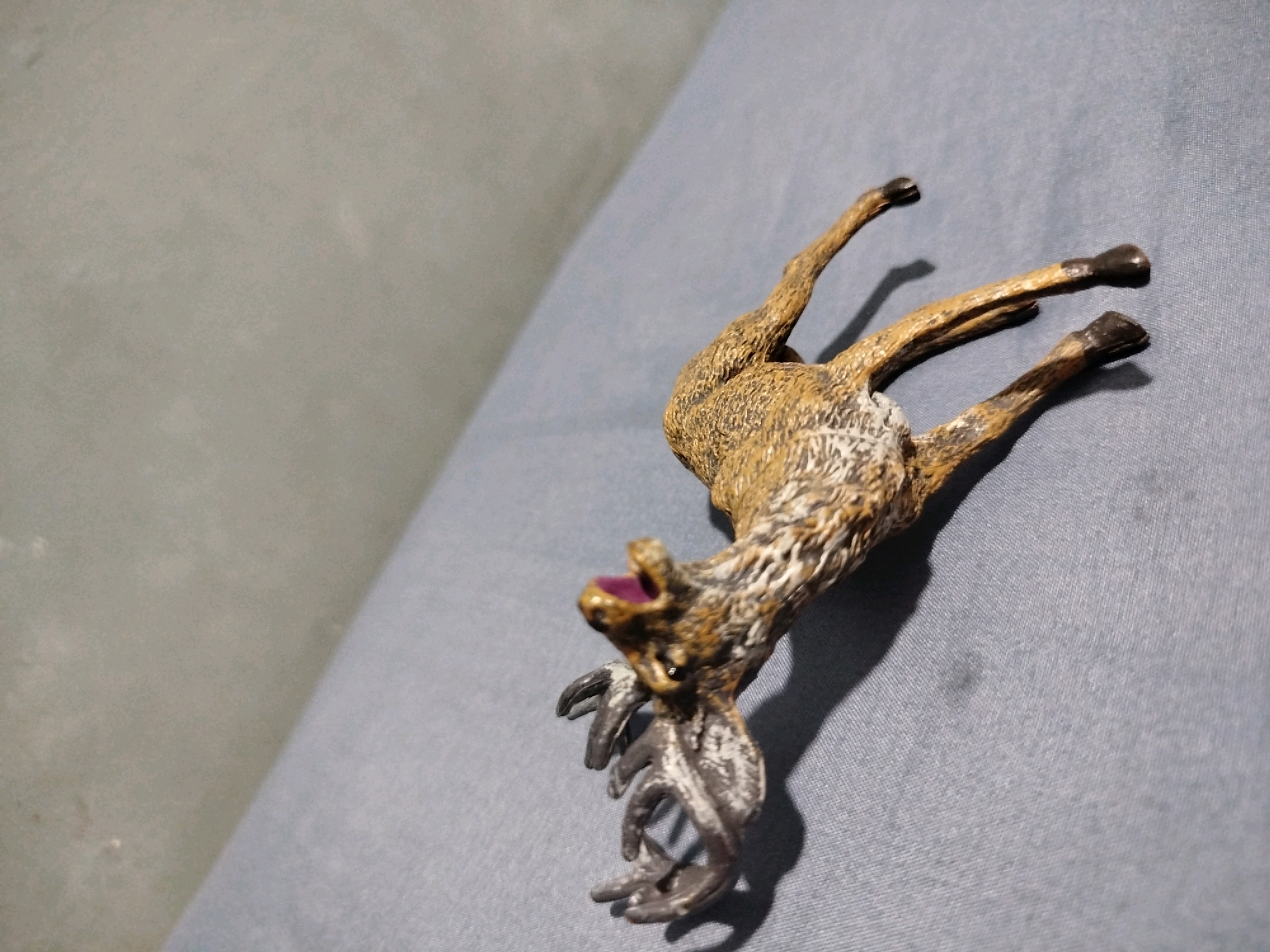 A plastic figurine of a deer is lying on its back on a textured, blue surface. The deer figurine is facing towards the left of the frame, with its legs splayed upwards. Its head is tilted back, and its mouth is open, revealing a purple interior. The figurine is detailed with a brown and white patterned coat and prominent antlers. The background consists of a gray, textured surface that takes up the left half of the image. There is no indication of the city of Mojoagung, Indonesia, within the image.