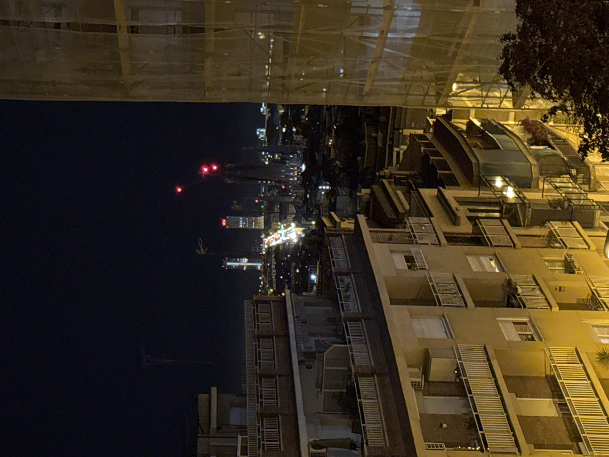 The image displays an urban cityscape at night in Barcelona, viewed from an elevated perspective. The sky is dark and clear, dotted with artificial lights from buildings and other sources.
In the mid-ground and foreground, a multi-story building, likely residential, is illuminated, showing numerous balconies equipped with white, horizontal louvered screens or blinds. One of the detailed crops reveals a person standing on a balcony, near a plant or object. The building's facade reflects a warm, yellowish light. Adjacent to this building, a lower roof area features what appear to be glass-covered structures or skylights, and some trees or greenery.
Towards the top of the frame (when correctly oriented), a large structure is covered by extensive scaffolding and protective netting, indicating ongoing construction or renovation.
In the background, a sprawling city skyline is visible, characterized by countless twinkling lights. Several distinct red lights are prominent against the dark sky, possibly originating from tall structures or distant aircraft. A faint, light-colored, hook-like shape is also discernible in the far distance.
The scene captures a quiet, contemplative urban night ambiance. No specific activities or events are taking place, apart from the general illumination of the city. No visible text is present in the image.