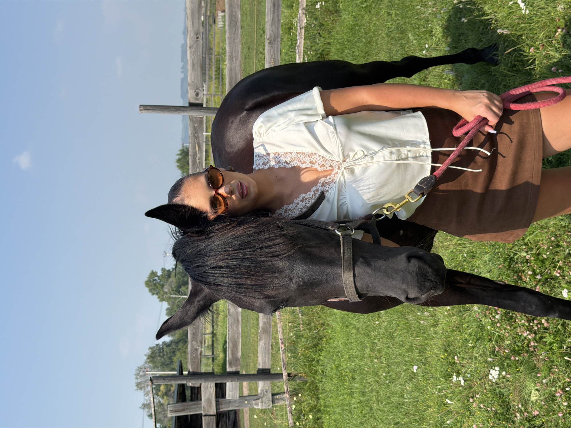 A young woman wearing sunglasses and a white lace-trimmed blouse with a brown skirt stands next to a black horse in a grassy field with a wooden fence in the background. The woman is holding a pink leash. The scene appears to be in the village of Esaulovo, Russia, under a clear blue sky.