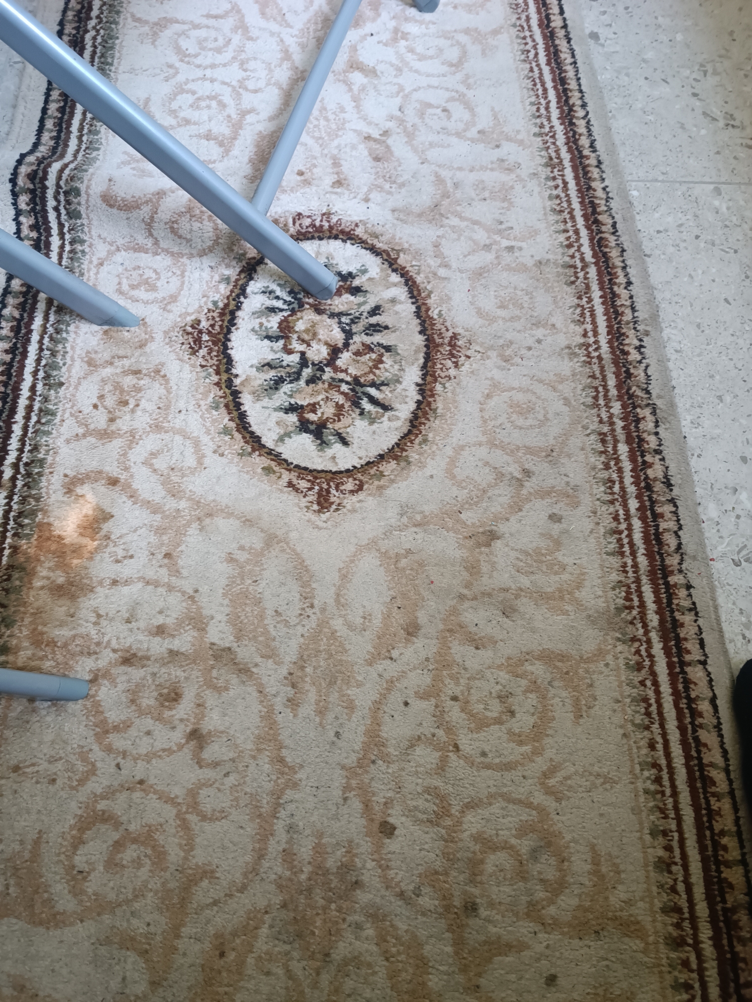 The image depicts a close-up view of a light-colored rug with a floral medallion pattern in its center. The rug is bordered by a strip of darker colors. Several light gray metal legs of a piece of furniture, possibly chairs or a table, rest on the rug. The rug appears to be placed on a speckled tile floor. The lighting suggests an indoor environment, potentially a home or a public space. There is no visible indication of the city of Burgas, Bulgaria.