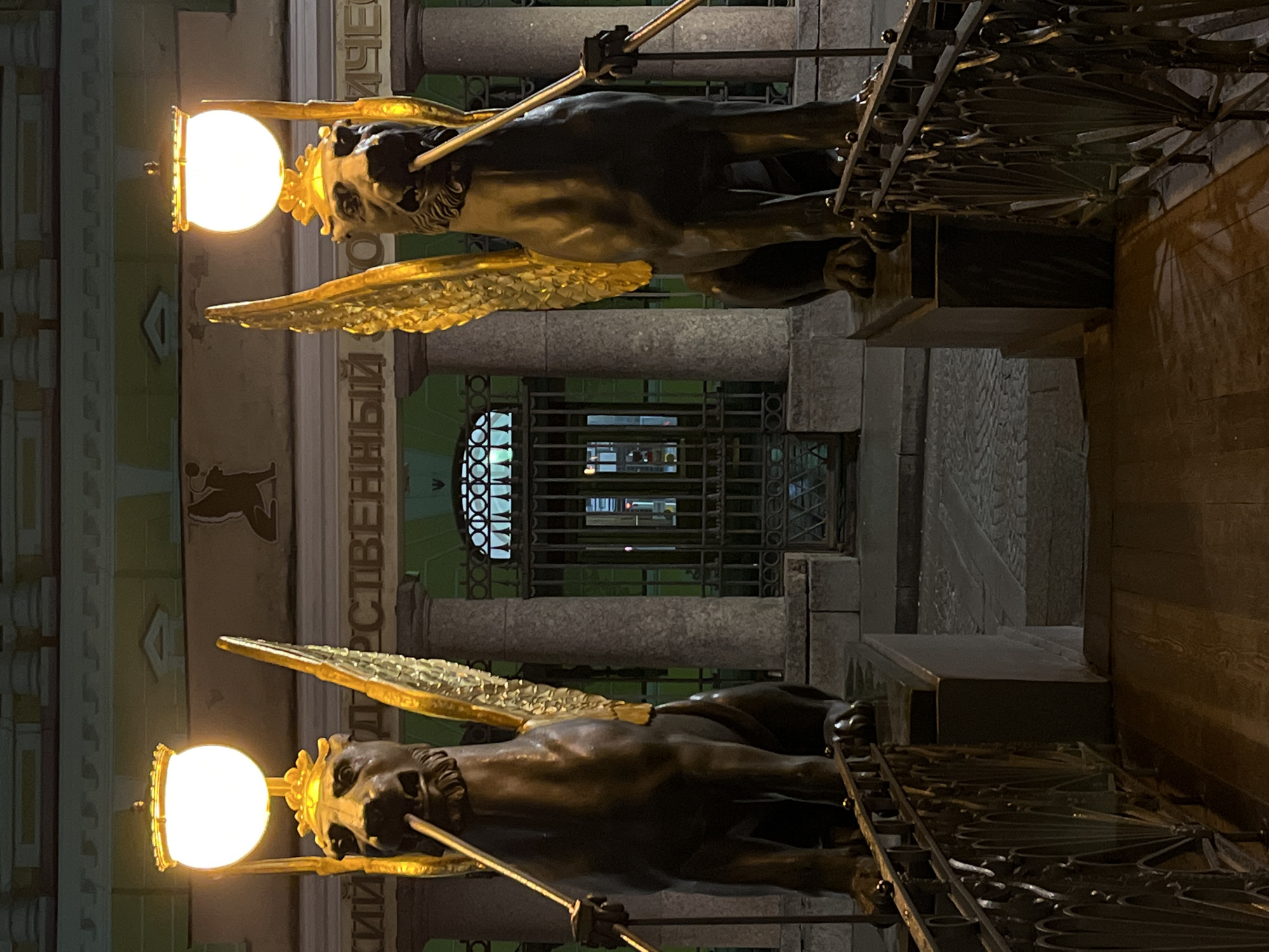 The image shows two winged lion statues holding light fixtures, located on a bridge in Saint Petersburg, Russia. The statues are dark bronze with golden wings. Each statue holds a lit spherical light fixture in front of its face and a staff. A wrought-iron fence with decorative elements is attached to the statues and forms part of the bridge's railing. In the background, there is a building façade with text in Cyrillic and architectural details such as columns and a gated entrance. The lighting suggests that the image was taken at night, with the light fixtures illuminating the statues and immediate surroundings. The photograph is rotated 90 degrees counter-clockwise.