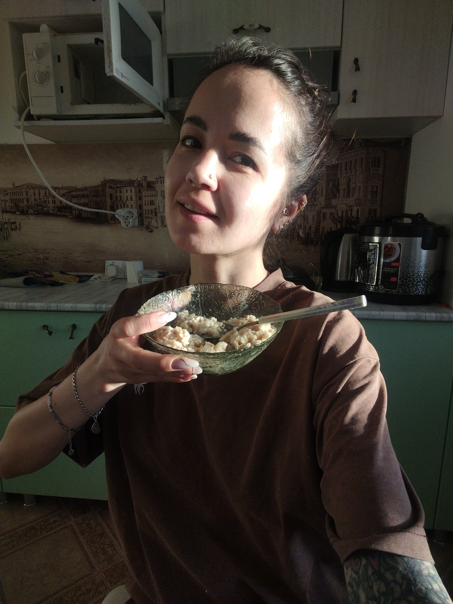 A woman is holding a bowl of porridge in her kitchen. She has dark hair pulled back, a nose ring, and ear piercings. She's wearing a brown t-shirt and has tattoos on her arm. Behind her are kitchen cabinets, a microwave, and a rice cooker. A picture of Venice is visible on the wall behind the counter. The location is in Noviy, Russia.