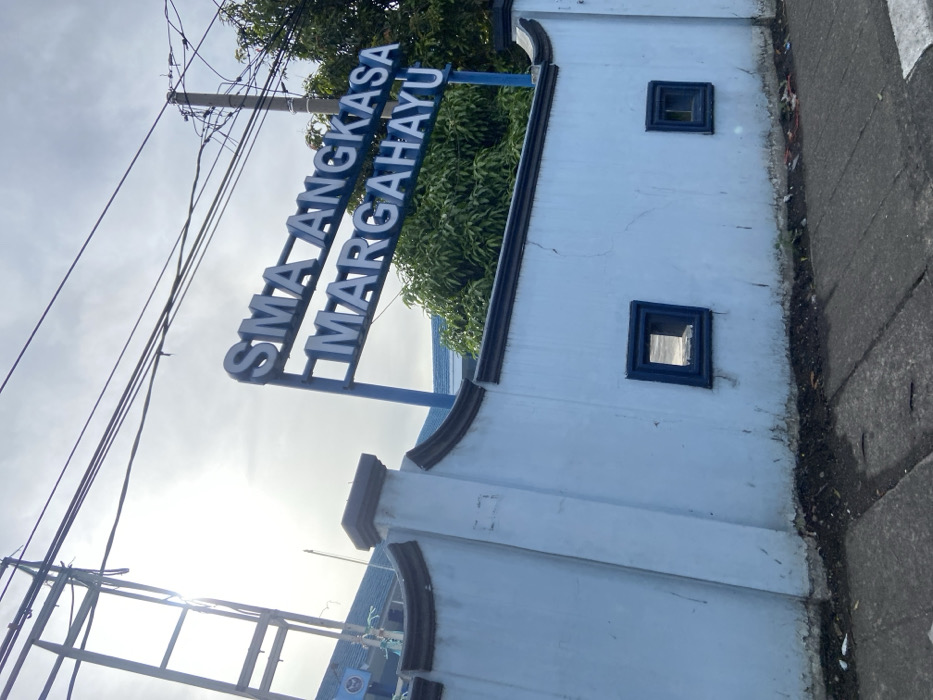 The image shows a blue and white wall with a sign that reads "SMA ANGKASA MARGAHAYU". The wall is located in Margahayu, Indonesia, at the coordinates -6.982, 107.563. The wall is topped with a brown molding and has two square, dark blue openings. There is a sidewalk in front of the wall, and several power lines are visible in the sky. The sky is overcast. The sign is mounted on a blue pole. There are no people visible in the image. The wall appears to be part of the entrance to the "SMA ANGKASA MARGAHAYU", which is likely a school.