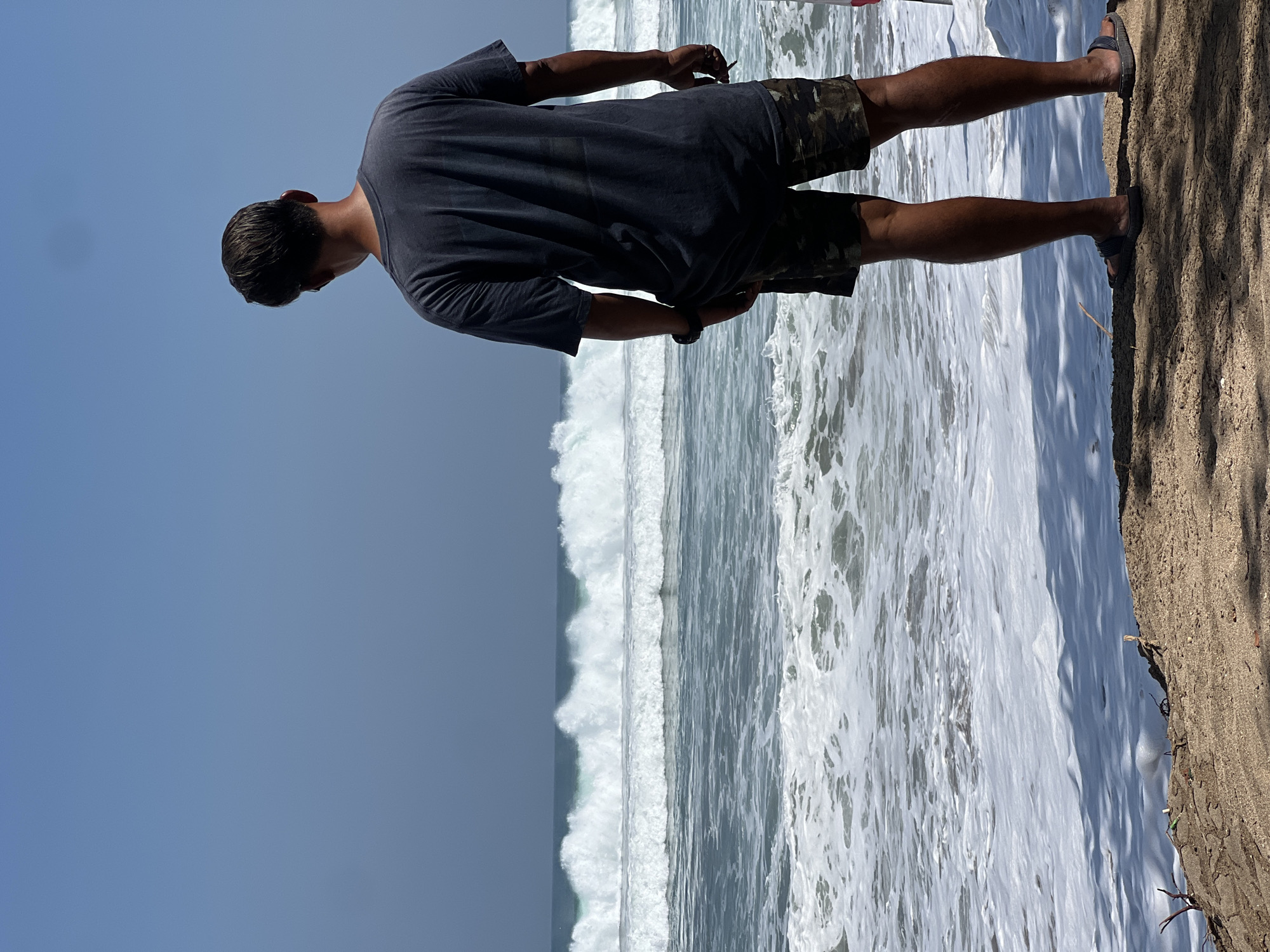 A man stands on a sandy shore, facing away from the camera, looking out at the ocean. He is wearing a dark gray t-shirt and camouflage shorts. The ocean is rough, with large waves crashing and white foam spreading across the shore. The sky is a clear blue. The scene is likely Kuta Beach, Bali, Indonesia, a popular tourist destination known for its strong waves and surfing conditions.