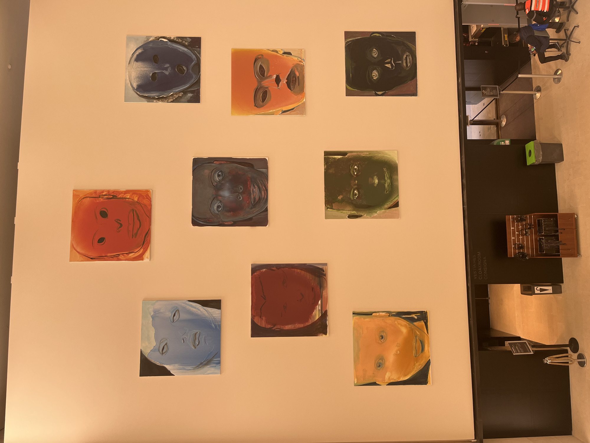 The image depicts a display of nine small, square portraits on a light-colored wall. The portraits are arranged in three rows and three columns, each featuring a face painted in varying, unnatural colors such as blue, orange, red, green, and black. The style appears expressive and somewhat distorted.
To the right of the wall is a black structure labeled "VESTIAIRES CLOAKROOM CONSIGNA" along with a logo of an umbrella. To the right of the black structure is what appears to be a security checkpoint with a metal detector and security personnel. The people are wearing uniforms or ID badges. The location appears to be in Paris, France due to the French language on the signage. The overall environment seems to be a museum or similar public space.