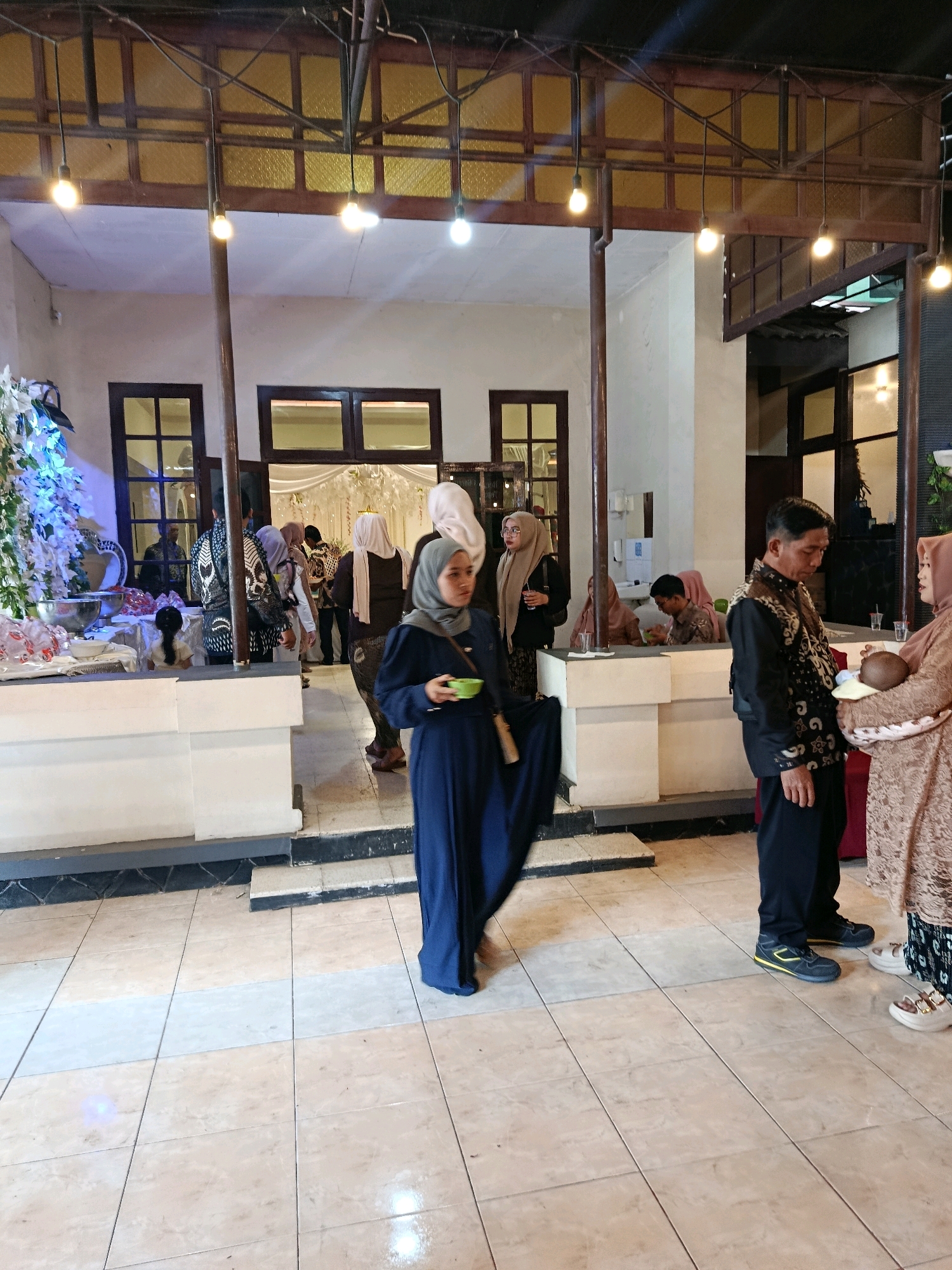 The image depicts an indoor event scene, likely a gathering or reception, at what appears to be a venue with an open-air pavilion-like structure. The architecture features wooden beams and latticework supporting a roof, with decorative lighting fixtures hanging down. The floor is tiled in a beige and cream pattern.
In the foreground, a woman wearing a dark blue hijab and dress is visible, holding a small green bowl. To her right, a man in a black patterned vest over a black shirt and dark pants stands next to a woman holding a baby. The woman holding the baby is wearing a light brown, ornate dress. Other individuals in traditional attire, including hijabs, are present in the background, some conversing and others standing near tables.
To the left, a floral arrangement with white and blue flowers is displayed on a table. The background shows doors and windows, with glimpses of more decorated spaces beyond. The overall ambiance suggests a formal or semi-formal social event.
The location, Tanggung, Indonesia, suggests a cultural context where such gatherings are common, potentially a wedding reception or community event. The style of clothing and the celebratory nature of the scene align with Indonesian cultural practices.