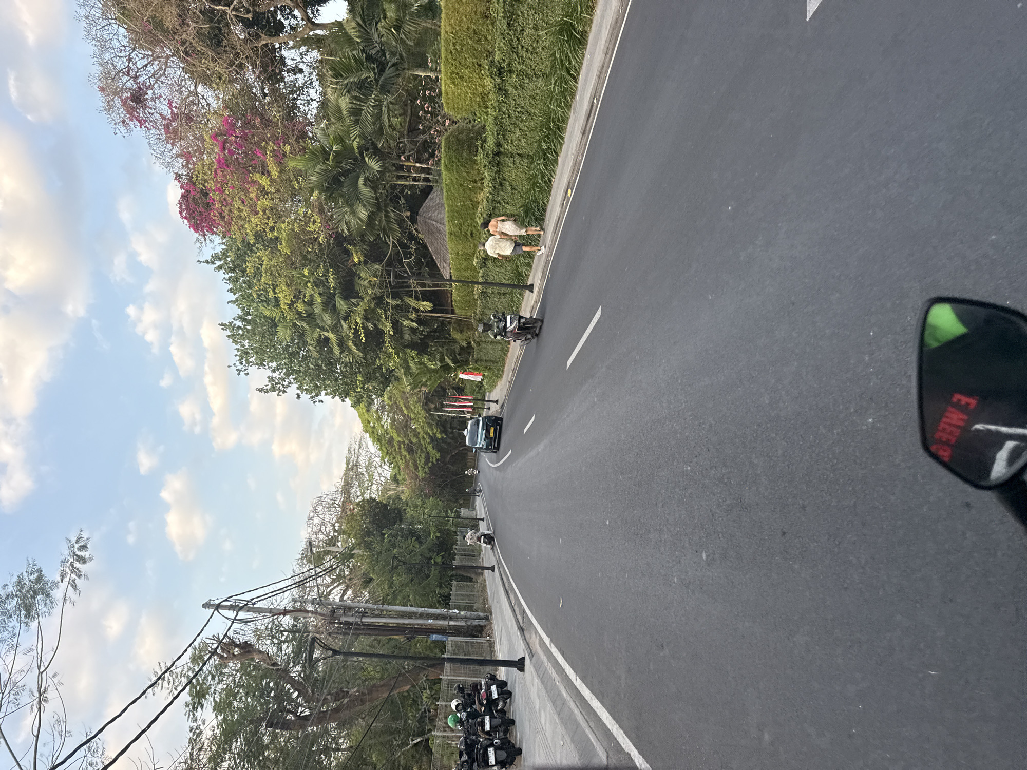 The image depicts a street scene in Denpasar, Indonesia, captured from the perspective of someone riding a motorcycle. The road is paved and curves to the right, bordered by a sidewalk and a well-maintained hedge. Several parked motorcycles with helmets are visible on the left side of the road, near a fence and utility poles. Further down the road, a green car is driving, and two people are walking on the sidewalk. The environment is lush with green trees and palm trees, with some flowering trees adding pops of pink to the foliage. The sky is partly cloudy, with a mix of blue sky and white clouds. The overall impression is one of a tropical urban environment.