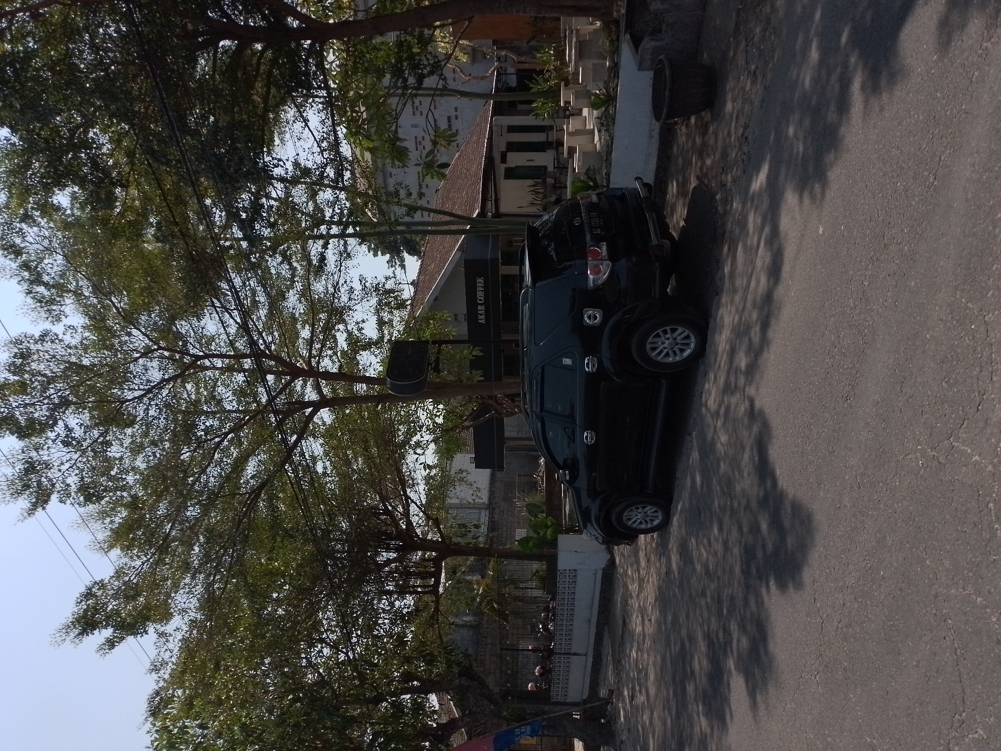 In the city of Wates, Indonesia, on a bright, clear day, a black SUV is parked on the side of a paved road. The SUV is positioned at an angle, facing towards the right of the frame. In the background, a building with a brown tiled roof and white walls is visible, with the sign "AKAR COFFEE" displayed prominently above the entrance. The area around the coffee shop appears to be an outdoor seating area with several cube-shaped stone tables and seats. Lush green trees with sparse foliage cast shadows across the road and the building. The environment suggests a typical street scene in a town or city, likely in a tropical climate given the vegetation. There are no visible people in the image, and the focus is on the car and the coffee shop.