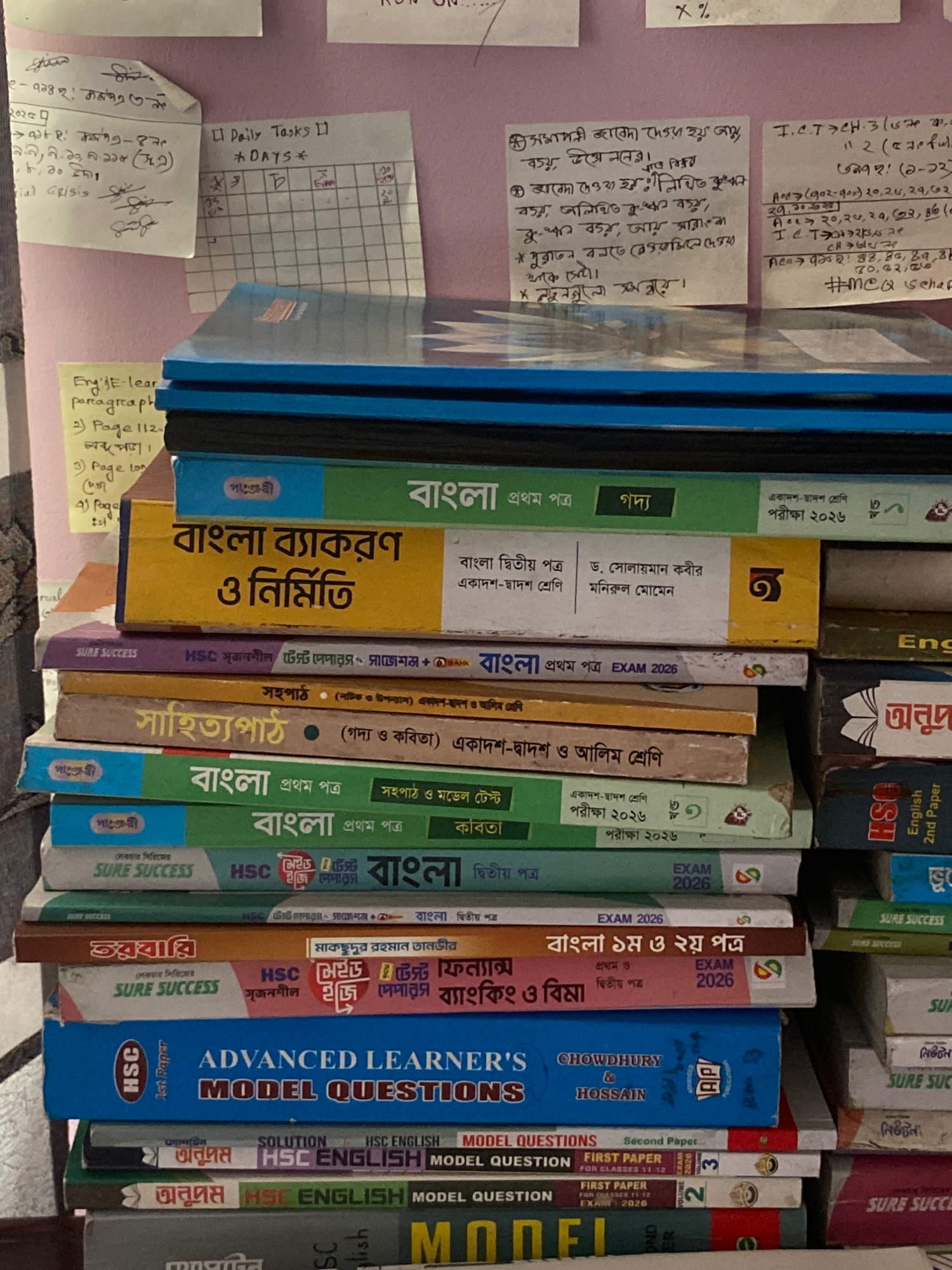 The image shows a stack of books, primarily textbooks and workbooks, on a surface. The books have titles and subjects in Bengali, such as "বাংলা ব্যাকরণ ও নির্মিতি" (Bengali Grammar and Construction), "সাহিত্যপাঠ" (Literature Study), and "বাংলা প্রথম পত্র" (Bengali First Paper). Some books also indicate "HSC" which refers to the Higher Secondary Certificate examination in Bangladesh.
Scattered around the books are various handwritten notes and papers. One note, written in Bengali, appears to be a daily task list or schedule with dates. Another note has "Daily Tasks" and "DAYS" written on it, with a grid underneath. There are also some smaller sticky notes with writing.
The background consists of a pinkish wall, suggesting an indoor setting. The overall impression is that of a study area or a collection of educational materials. The books are neatly stacked, implying organization.
Based on the presence of HSC examination-related materials and common subjects taught in Bangladeshi schools, this scene is likely located in a residential or educational setting within Bangladesh. Without further information specific to the location of Hemayetpur, Bangladesh, it is not possible to pinpoint the exact location within the city. However, the visual cues strongly suggest a Bangladeshi context. There are no people or actions depicted in the image.