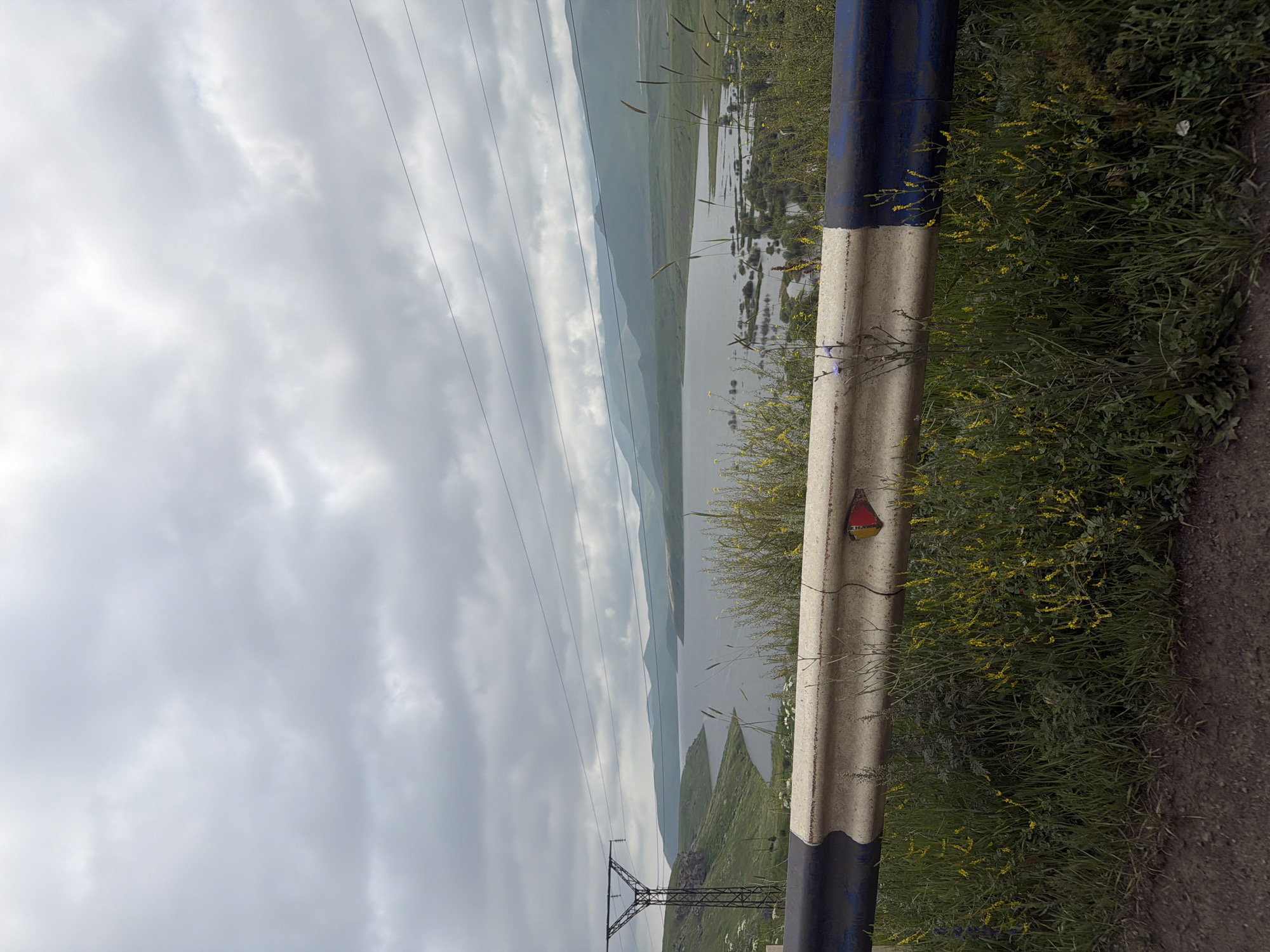 The image depicts a scenic landscape with a body of water, rolling hills, and a cloudy sky. In the foreground, there is a roadside guardrail. To the left, a large power line tower is visible. The environment suggests a rural or mountainous area. The location is identified as Tsghuk, Armenia.