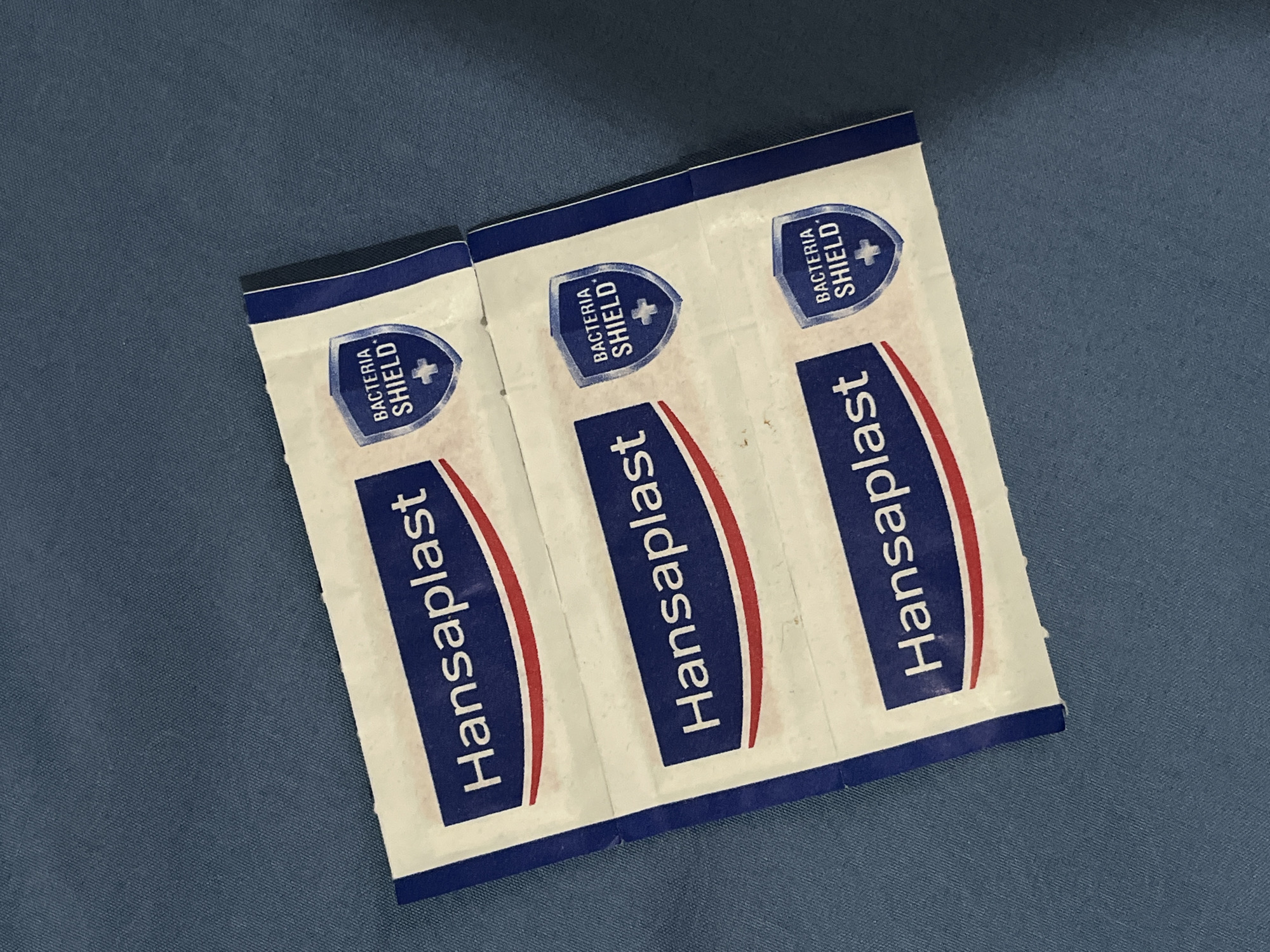 Three individually wrapped Hansaplast bandages with "Bacteria Shield" branding are arranged diagonally on a textured blue fabric surface. The bandages are stacked one above the other, with slight overlap. The fabric appears to be a dark, muted blue. No people are present in the image. There is no information in the image to indicate the location is Teluk Air, Indonesia.
