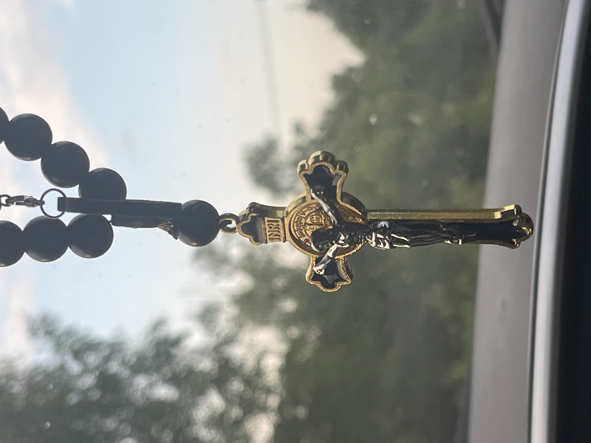 The image shows a close-up of a crucifix pendant hanging from a string of dark beads, likely a rosary. The crucifix itself is detailed, with a depiction of Jesus Christ and the inscription "INRI" at the top. The pendant has a metallic sheen, appearing to be gold and black. The background is blurred, showing greenery and a sky with soft clouds, suggesting an outdoor setting, possibly viewed through a window. The foreground is dark and out of focus, hinting at the interior of a vehicle. The location of Никольское, Russia is not identifiable from the visual information provided in the image.