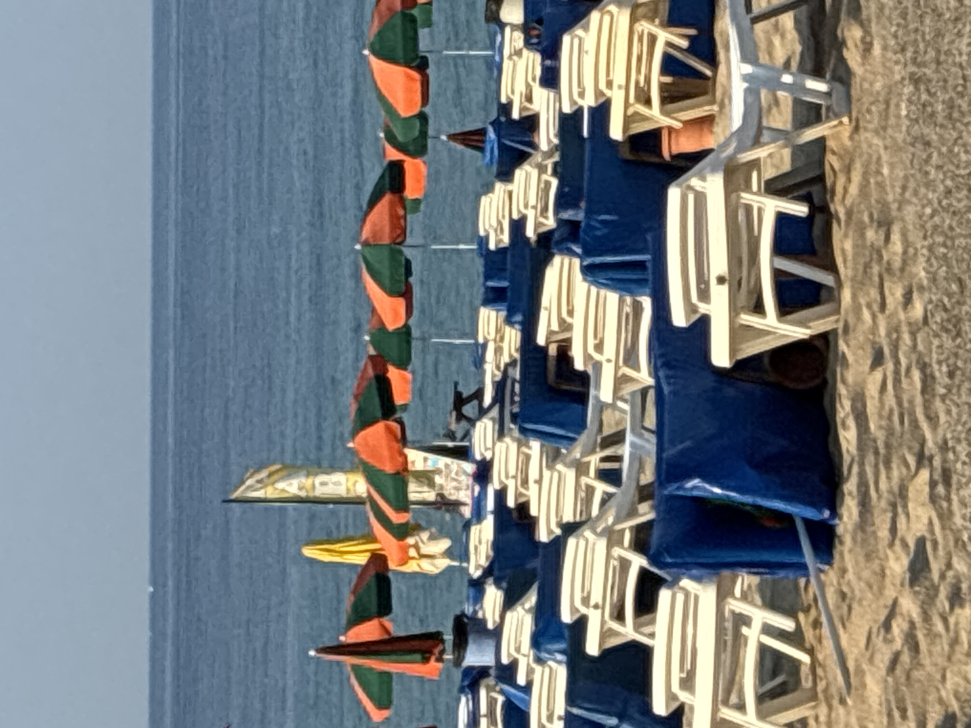 The image depicts a beach in Alanya, Turkey, with rows of empty lounge chairs and umbrellas set up on the sand. The sea is visible in the background, with a clear blue sky above. The lounge chairs are mostly covered with blue tarps, and several open umbrellas with orange and green stripes are spread across the scene. A few flags are also visible, one with yellow and white stripes. The sand is light brown and textured, with some scattered debris. The overall impression is a quiet, possibly early morning or late evening scene, as there are no people present on the beach.