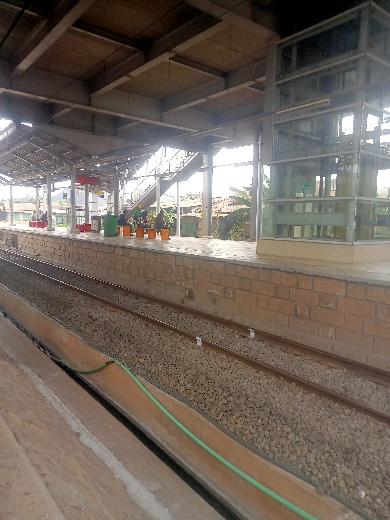 A vertical medium shot captures a train station platform in Unknown, Unknown. The platform is constructed with brickwork at the base and concrete on top, with two sets of train tracks running alongside. Several people are visible on the platform, some seated on orange benches and others standing. A green trash bin is also present. To the right, a modern glass elevator enclosure stands out. In the background, buildings and vegetation suggest an urban or semi-urban environment. The scene appears to be outdoors, with overhead shelter provided by the station's roof structure.