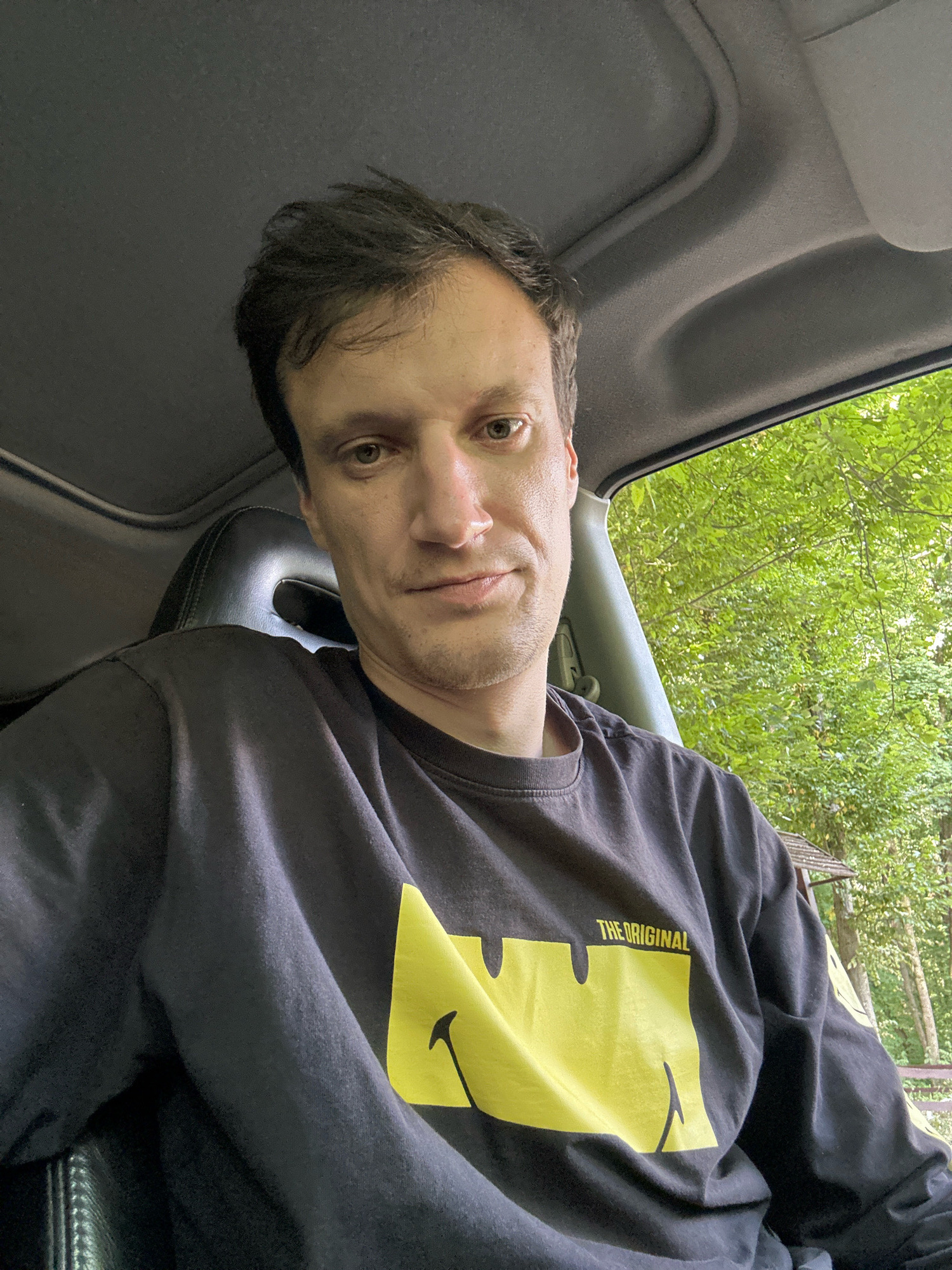 A man is sitting in the driver's seat of a vehicle, looking at the camera. He has short, dark hair and light-colored eyes. He is wearing a black long-sleeved shirt with a large yellow graphic on the front that says "THE ORIGINAL" above a smiley face. The interior of the car is visible, including a black leather seat and the roof. Outside the car window, lush green trees and foliage are visible, suggesting an outdoor, possibly wooded, environment. The lighting suggests it is daytime. The location is not discernible from the image.