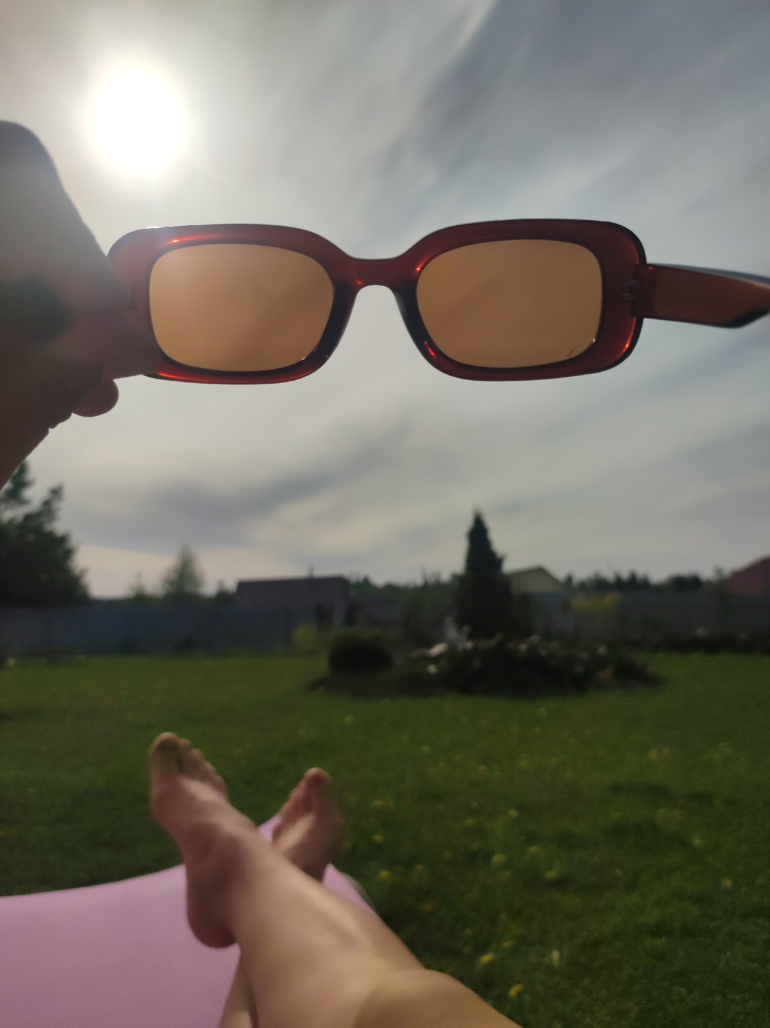 A person is relaxing on a pink surface in a grassy yard with their bare feet visible in the foreground. The person is holding brown-rimmed sunglasses up to the sky, partially obscuring the view. The sky is overcast with the sun visible behind the glasses. In the background, there is a green lawn with dandelions, a dark fence, trees, bushes, and houses. The location is possibly Фоки, Russia.