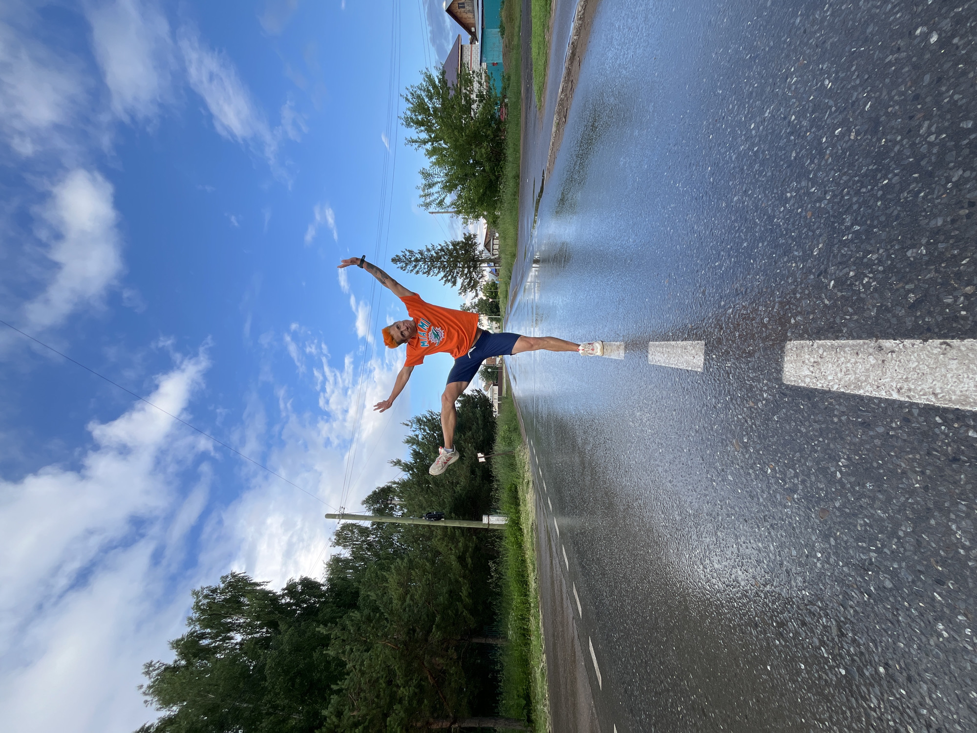 A young man with bright orange hair is balancing on one leg in the middle of a wet, asphalt road, his arms outstretched in a dynamic pose. He is wearing an orange t-shirt with a Miami Dolphins logo and dark blue shorts. The road appears to have recently experienced rain, with puddles reflecting the overcast sky and surrounding trees. The scene is set in a rural or suburban area, indicated by the presence of trees, power lines, and a few houses in the background. This appears to be a public road, possibly in Chaikovsky, Russia, given the context.