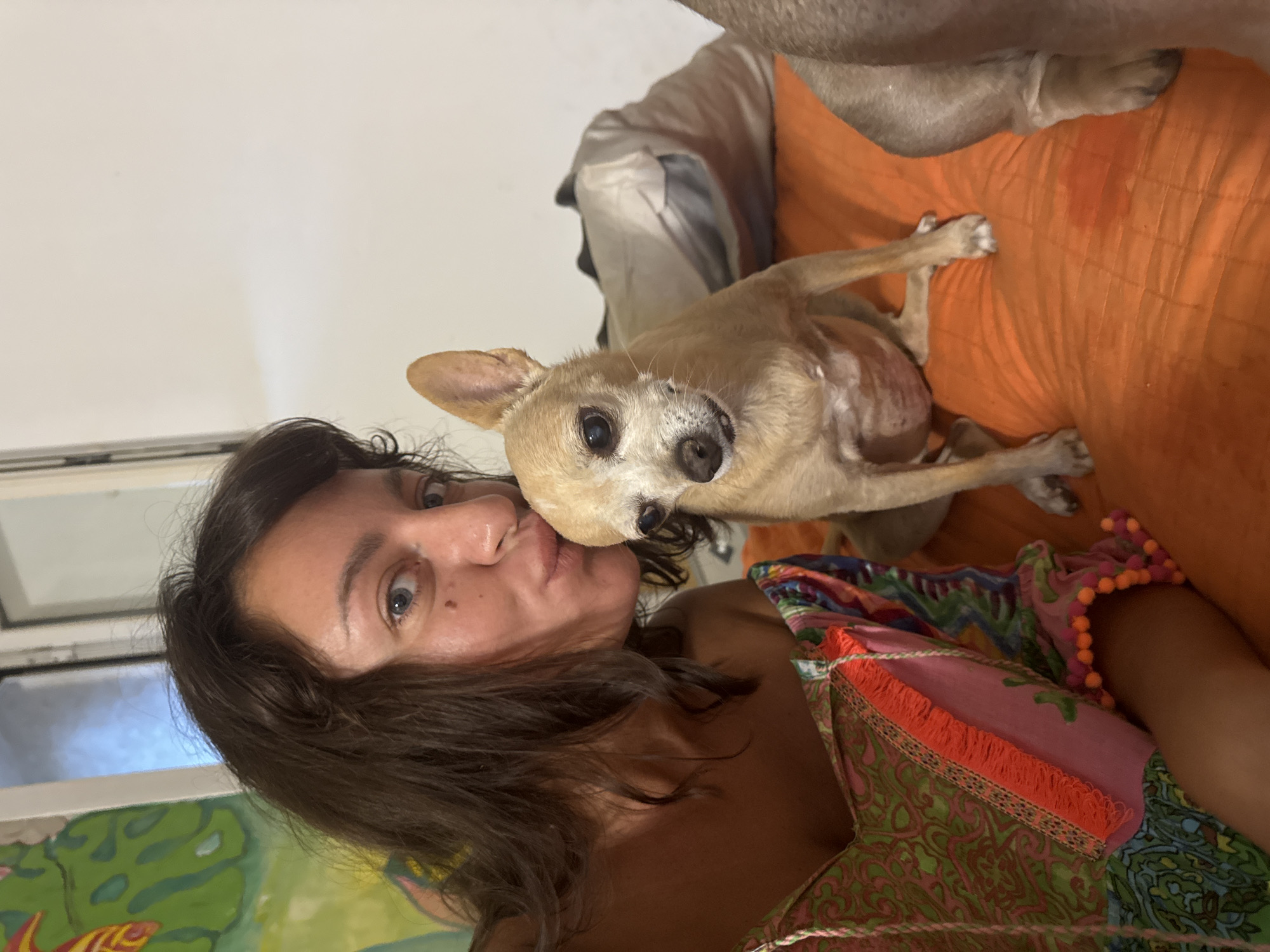 A woman and a small, tan Chihuahua are seen in a close-up, playful pose. The woman is on the left, her face turned towards the camera with her lips puckered as if to kiss the dog, who is leaning in towards her. The dog has large, dark eyes and its head is tilted, looking directly at the viewer. The woman has dark, wavy hair and her eyes are wide open. She is wearing a colorful, patterned garment with fringed and beaded embellishments. The background appears to be indoors, with an orange-patterned surface behind the subjects, possibly a couch or cushion. To the left, a portion of a window or framed artwork with green and yellow abstract designs is visible. The overall mood is intimate and affectionate. The lighting suggests it could be daytime, as there's ambient light coming from the left, illuminating the scene. The image is oriented vertically, with the subjects appearing to be on their side. There is no visible text in the image.