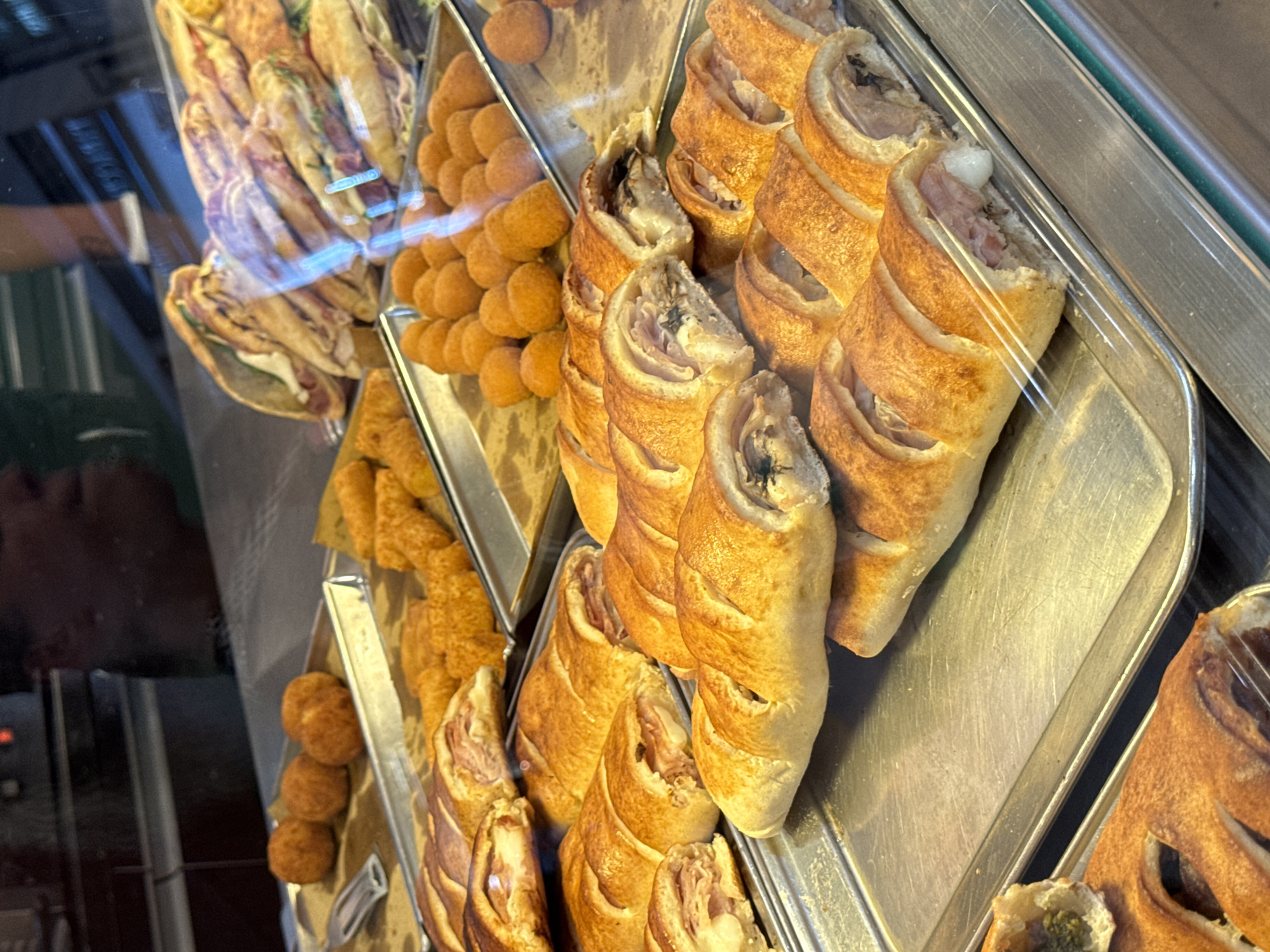 The image shows a display case filled with various fried and baked goods, likely in a bakery or food stall in Palermo, Italy. The foreground features several long, golden-brown baked pastries, sliced open to reveal fillings of ham and cheese, and possibly mushrooms or herbs. Behind these, on a separate tray, are numerous small, round, deep-fried items, possibly arancini or croquettes. Further back, there are sliced sandwiches or calzones with visible fillings of meat and greens. The display is set within a glass enclosure, reflecting the surroundings and a blurred figure. The overall impression is a vibrant and abundant offering of local street food or bakery items.