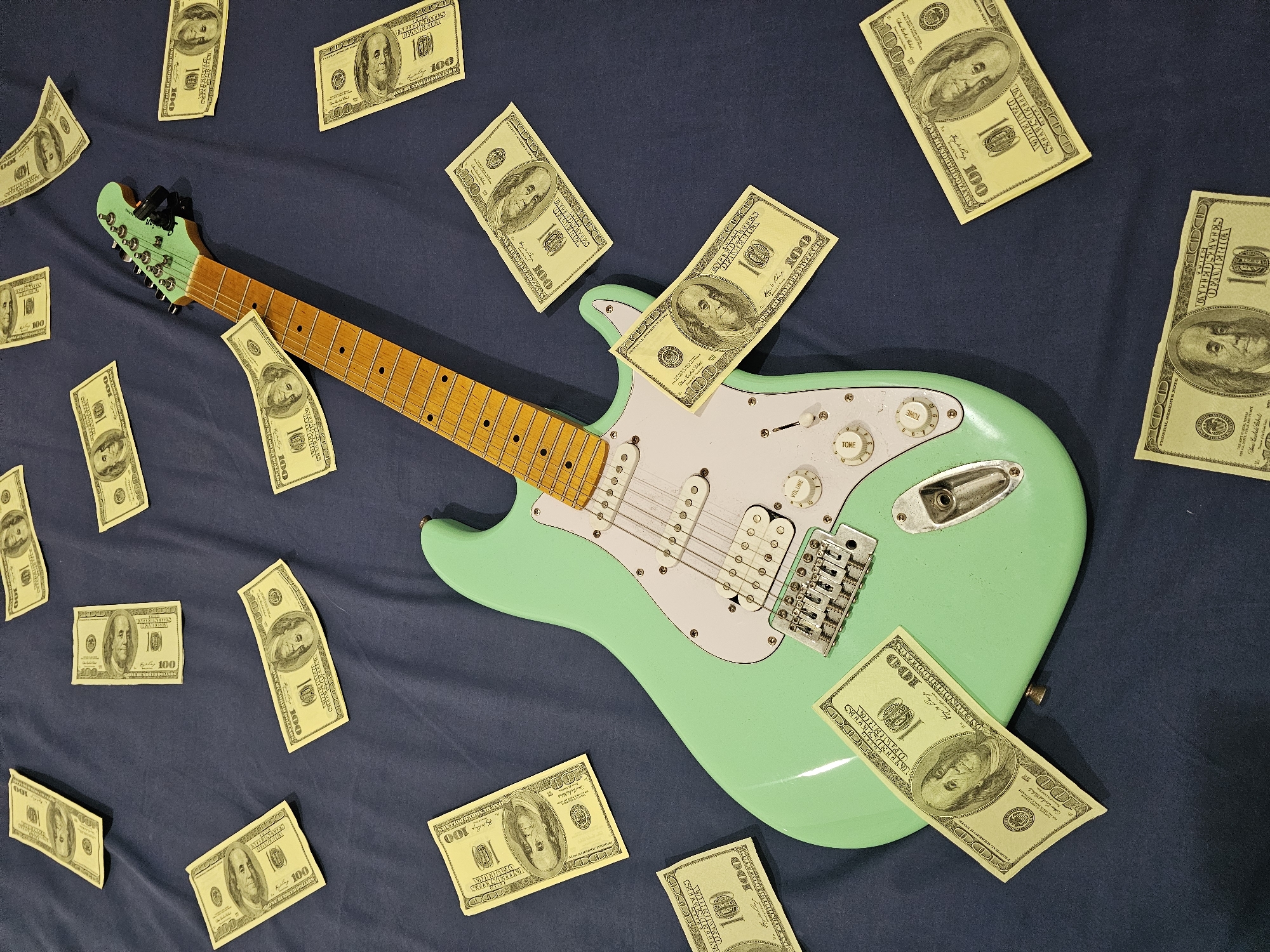 The image shows a light green electric guitar lying on a dark blue fabric surface, surrounded by scattered US hundred-dollar bills. The guitar is positioned diagonally in the center of the frame. The currency is distributed around the guitar. The image was not taken in Denpasar, Indonesia, there is no evidence in the image to suggest it was.