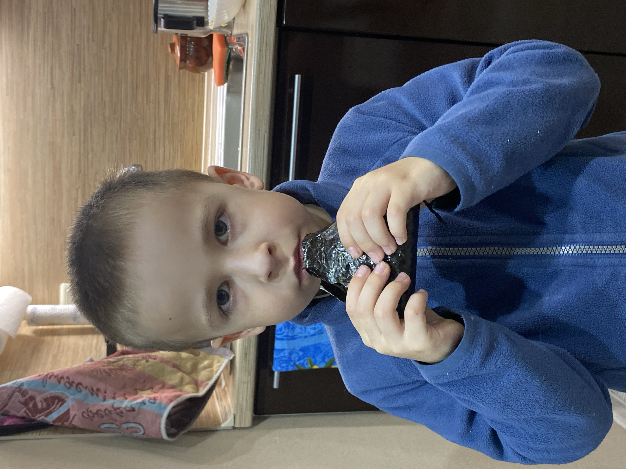 A child wearing a blue fleece jacket is eating a dark, possibly seaweed-wrapped food item. The setting appears to be a kitchen, with wood-patterned walls and cabinetry visible in the background. A towel and other kitchen items are also present. There is no definitive evidence to confirm this location as Markovskii, Russia.
