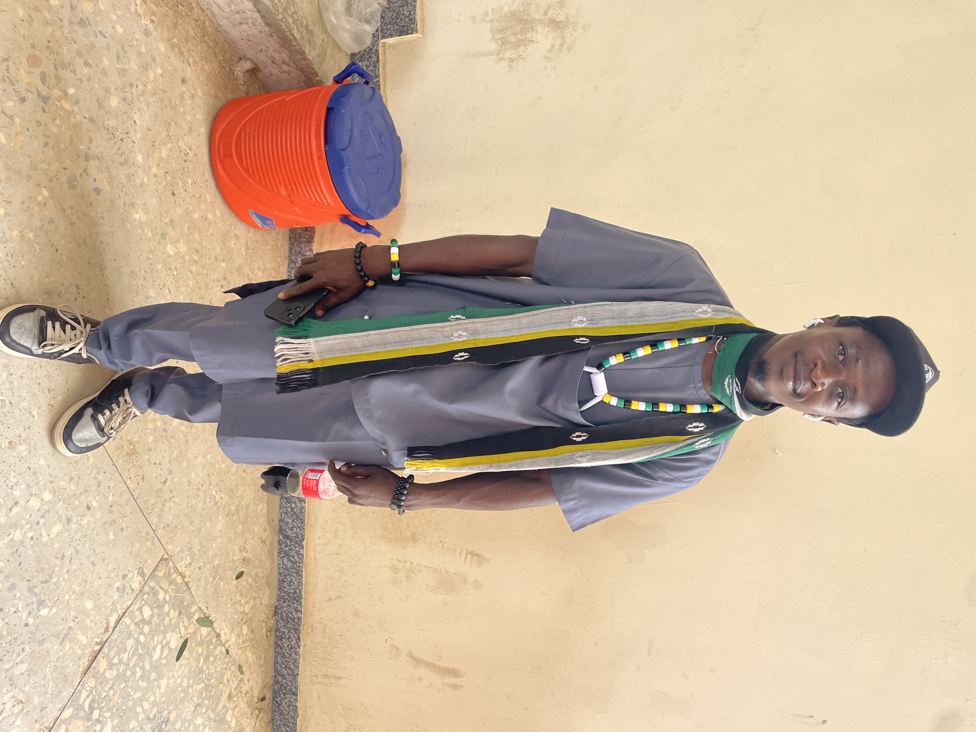 A medium shot captures a man standing in front of a beige wall. He is wearing a black baseball cap, a grey outfit consisting of a loose-fitting shirt and pants, a green bandana around his neck, and a colorful scarf with black, white, yellow, and green stripes. He also sports beaded necklaces and bracelets. In his right hand, he holds a red-capped bottle of soda, and in his left hand, he holds a black smartphone. To his left is an orange and blue cooler. The ground is covered in a terrazzo-like material, with scattered green leaves. The lighting suggests it is daytime.