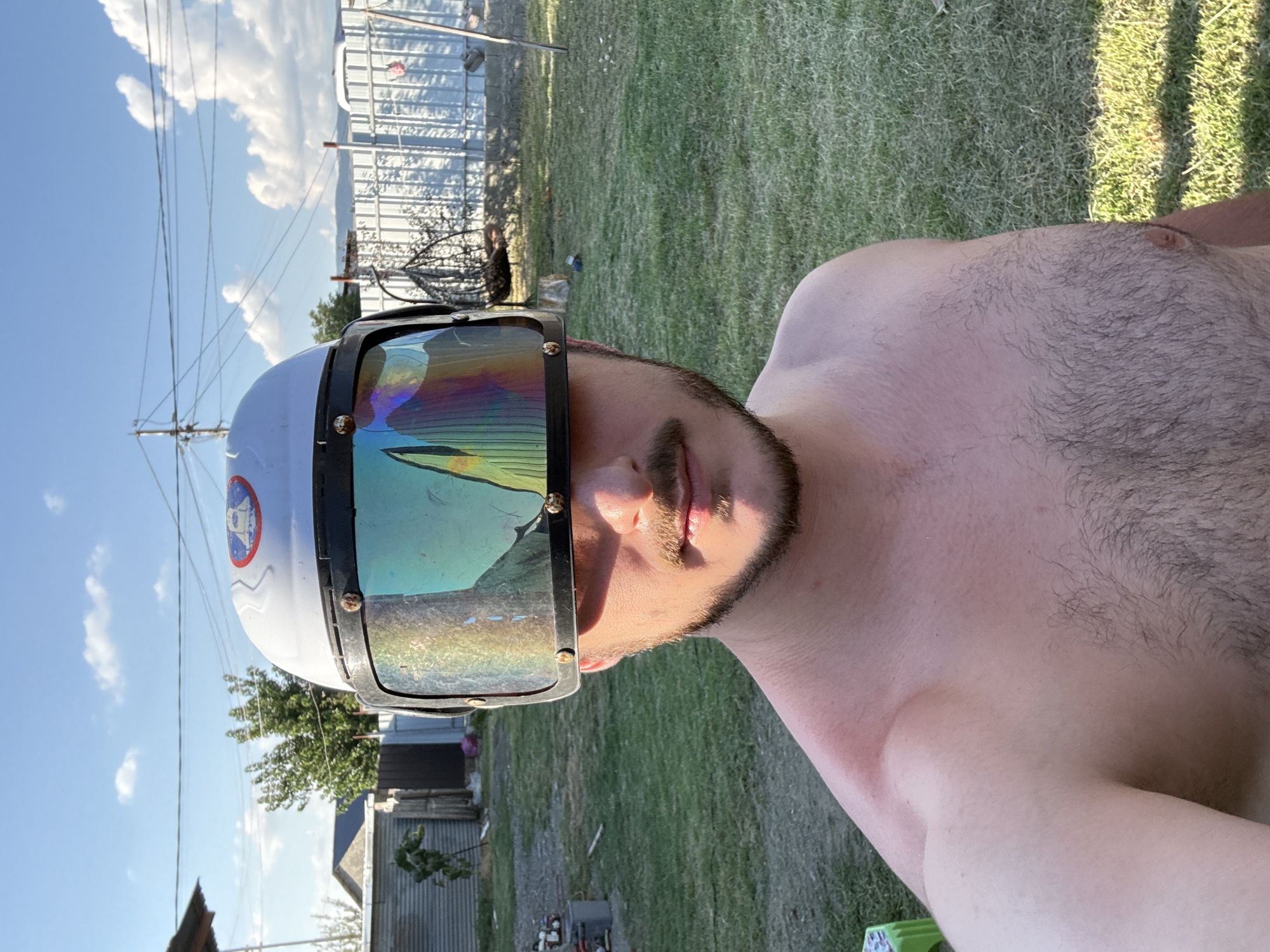 A man with a beard and mustache, shirtless, stands outdoors, wearing a white helmet with a large, reflective visor that displays a rainbow of colors. The background consists of a grassy yard with a white fence, some trees, and a clear blue sky with white clouds. The scene appears to be a residential area, possibly in Tbilisi, Georgia, given the architectural style of the nearby buildings. The lighting suggests it is daytime, likely late afternoon or early evening due to the warm light. The man's skin shows a slight redness on his shoulder, indicating potential sun exposure.