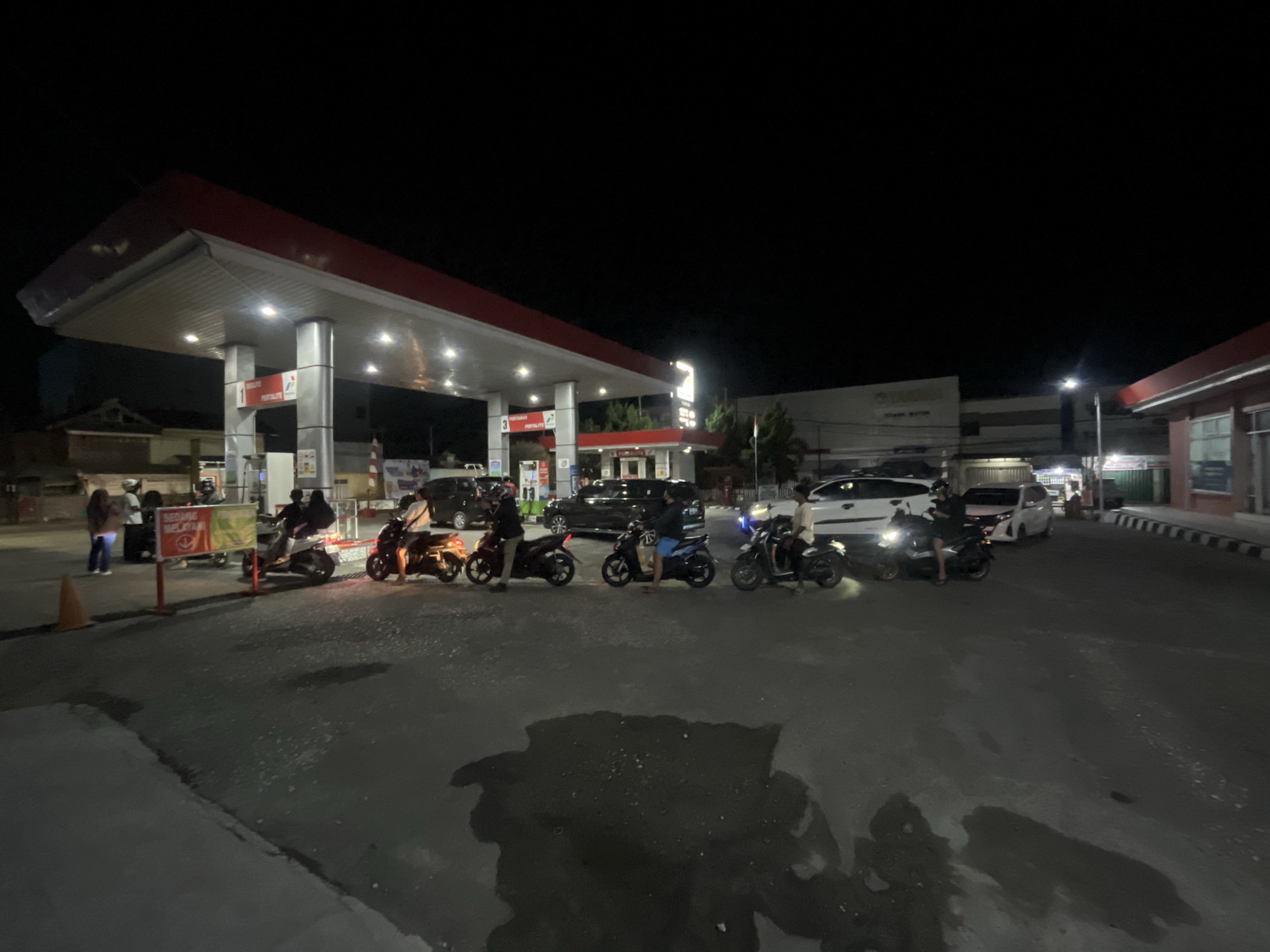 At night, a gas station in Luwuk, Indonesia, is illuminated by artificial lights. A red-and-white canopy extends over several fuel pumps, with one pump displaying the number "1" and another the number "3". Several motorcycles and cars are present, some of which appear to be queuing at the pumps or waiting. One person is visible standing at a pump, and others are on motorcycles. A sign with text and an orange traffic cone are also present. The ground is paved and appears wet in places, reflecting the lights. Trees and buildings are visible in the background.