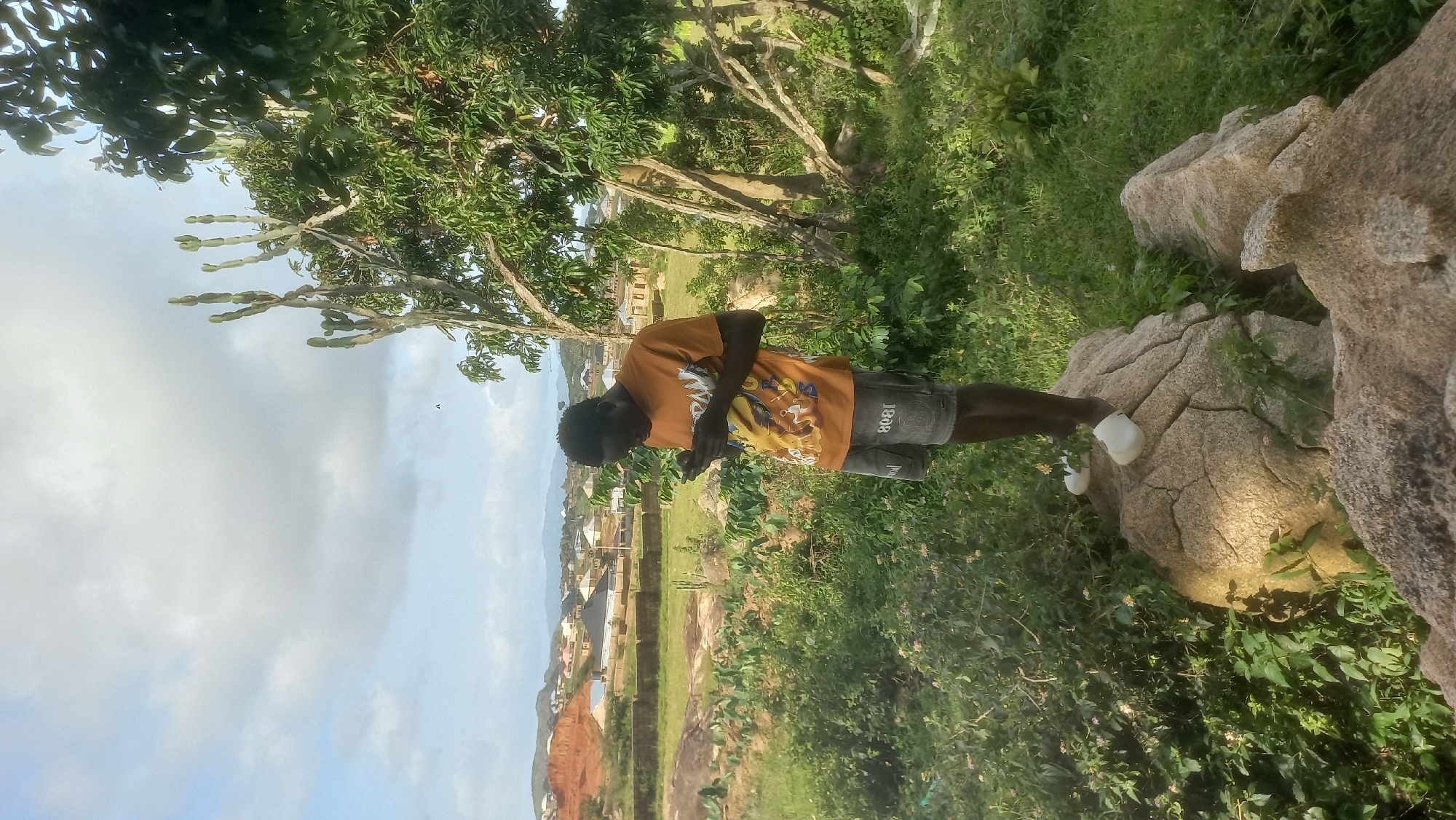 A young Black man stands on a large rock formation, looking down at a mobile phone in his hands. He is wearing an orange t-shirt with a graphic design and grey shorts. He has on white slip-on shoes. The man is surrounded by lush green vegetation. In the background, a village is visible nestled amongst rolling hills. The village appears to be a collection of houses with red-tiled roofs. The sky above is partly cloudy with a mix of blue sky and white clouds. The overall scene depicts an outdoor setting in a rural or semi-rural area. The location is identified as Kwang, Nigeria.
