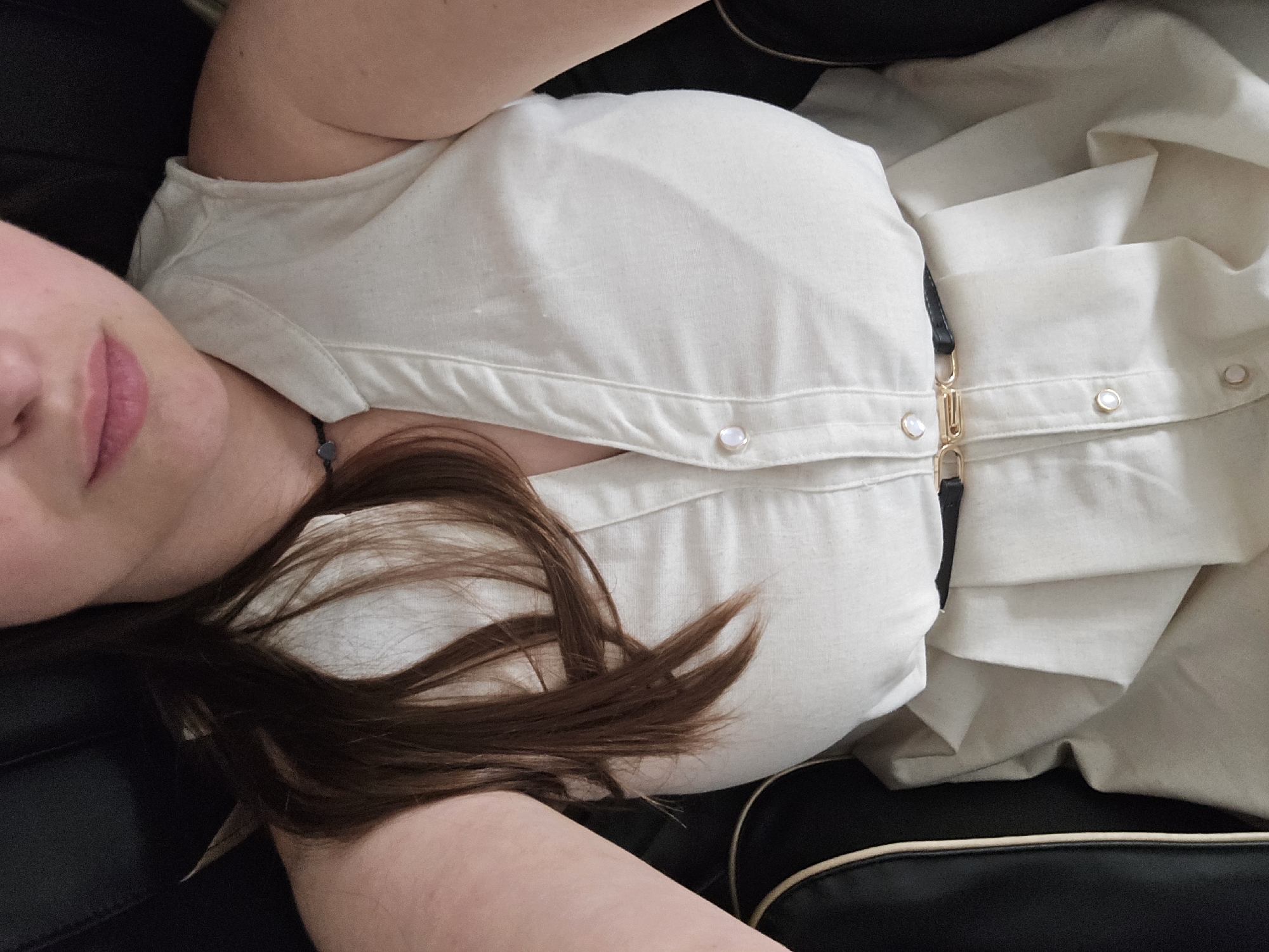 The media file is a close-up, overhead shot of a person lying down, viewed from a top-down perspective. The person has fair skin and is wearing a light-colored, button-down top. Their lips, colored with pink lipstick, are partially visible, along with strands of brown hair. The person appears to be resting on a black surface, possibly a piece of furniture or upholstery, with tan trim visible. The lighting suggests an indoor setting. There is no visible text in the image. The image does not provide enough information to determine the time of day, weather, or specific emotions. Given the location context of Kyiv, Ukraine, the scene is set within this city.