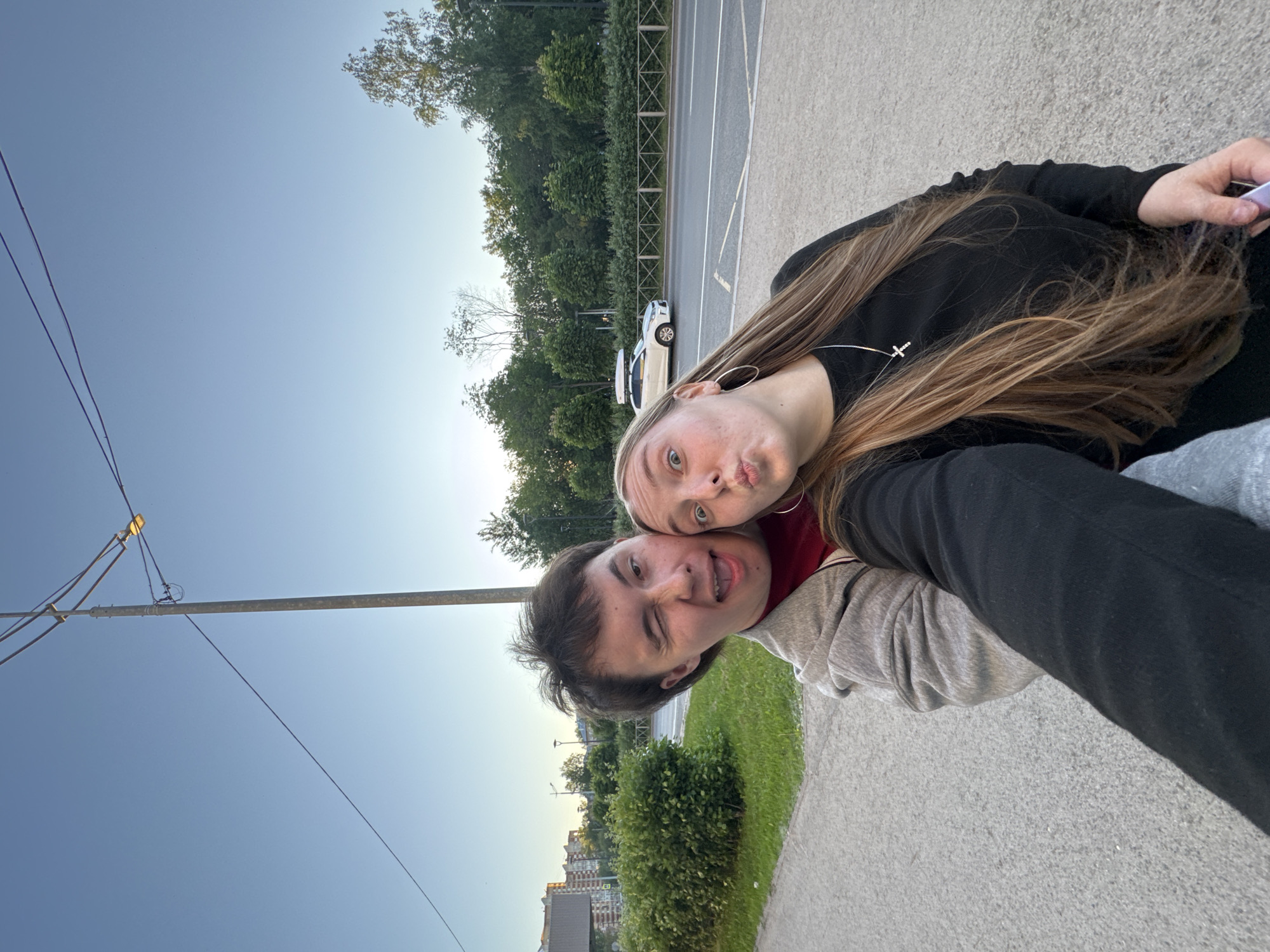 A young man and a young woman are taking a selfie outdoors in an urban environment. The man is on the left, winking and sticking his tongue out. He is wearing a grey hooded jacket over a maroon t-shirt. The woman is on the right, puckering her lips. She has long, blonde hair and is wearing a black long-sleeved shirt with a silver cross necklace. They are close together, suggesting they are a couple.
In the background, there is a sidewalk, a grassy area with bushes, a road with a white car, and trees. A tall utility pole with wires and a streetlight is visible on the right. The sky is clear and blue, indicating it is daytime. The buildings in the far background suggest an urban setting. The presence of buildings and a road with a car indicates they are in a city. Given the typical architecture and vegetation, the scene is consistent with a location in Perm, Russia.