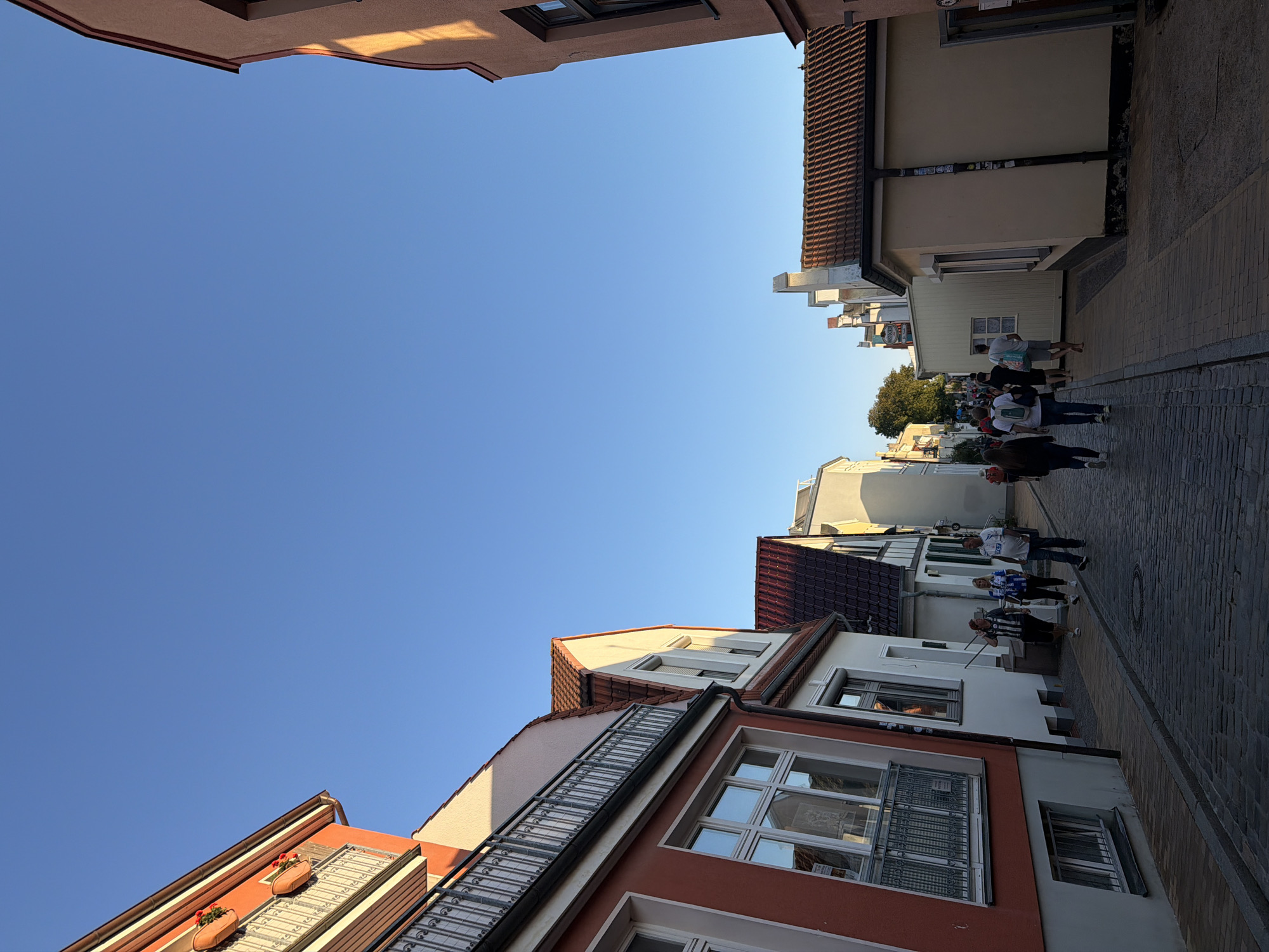 This is a sunny day photograph of a narrow cobblestone street in the historic district of Rostock, Germany, likely near the harbor area given the traditional architecture. On the left, a building with a vibrant orange facade and white trim features a balcony with a decorative metal railing and flower boxes. To its right, a white building with a tiled red roof and a modern window is visible. The street is lined with several other buildings, most with tiled roofs, in varying shades of white, yellow, and red.
Several pedestrians are walking along the street. In the foreground, on the right sidewalk, a man and a woman wearing matching blue and white patterned shirts are walking away from the camera. Further down the street, a group of people are walking in both directions, some carrying bags. The sky is a clear, bright blue, indicating good weather. The lighting suggests it is late afternoon or early evening, casting long shadows and highlighting the warm tones of the buildings. The scene depicts a lively, tourist-friendly atmosphere.