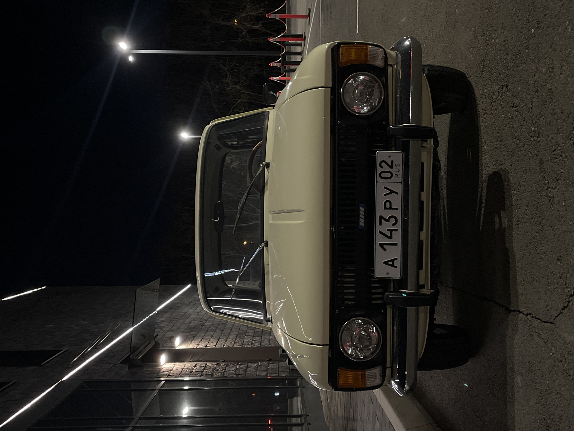 The image shows the front of a light-colored car, likely an older model, parked on a street at night. The license plate reads "A 143 PY 02 RUS," indicating the car is registered in Russia. The presence of streetlights and a modern building with illuminated linear lighting suggests an urban location. The exact location within Krasnoyarsk cannot be determined from the image alone.
