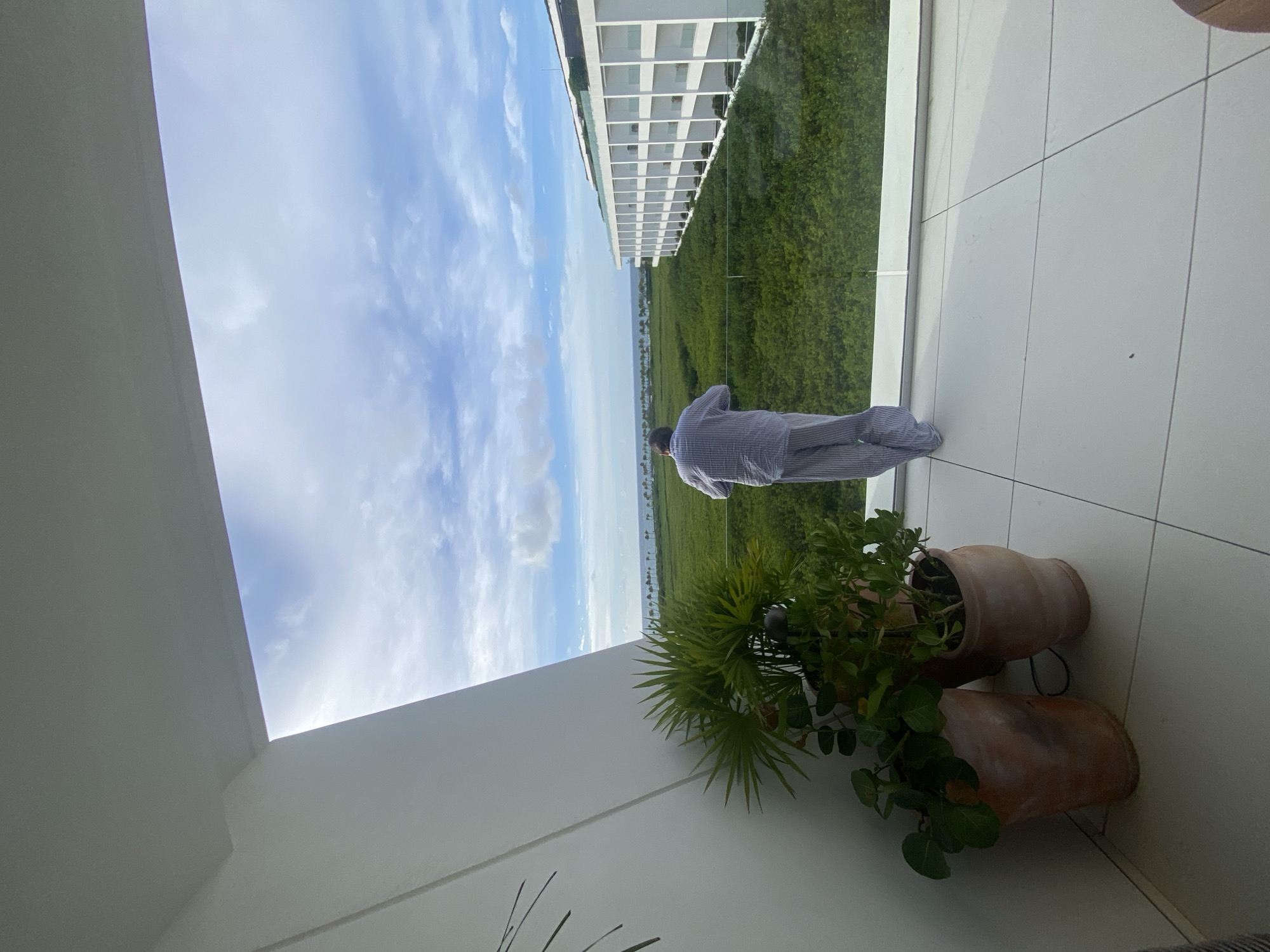 A man in pajamas is standing on a white tiled balcony overlooking a green landscape that may be a mangrove or lagoon. The man's back is to the viewer. In the foreground are two potted plants. In the distance are buildings along the coast. The view is from El Caracol, Mexico.