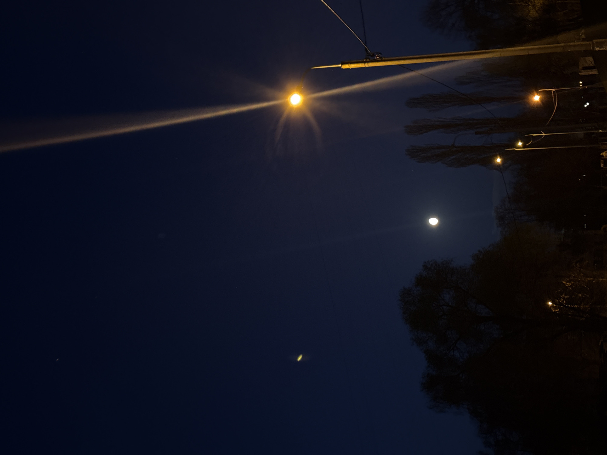 The image shows a night scene in Kyiv, Ukraine. A bright street light shines prominently from the upper right, casting a strong beam across the dark blue sky. Below the street light, another light source, possibly the moon, is visible. Trees line the right side of the frame, their silhouettes dark against the sky. Another, elongated light source is visible below the moon. Electrical wires extend from the upper right corner. Other light sources are visible from the trees.