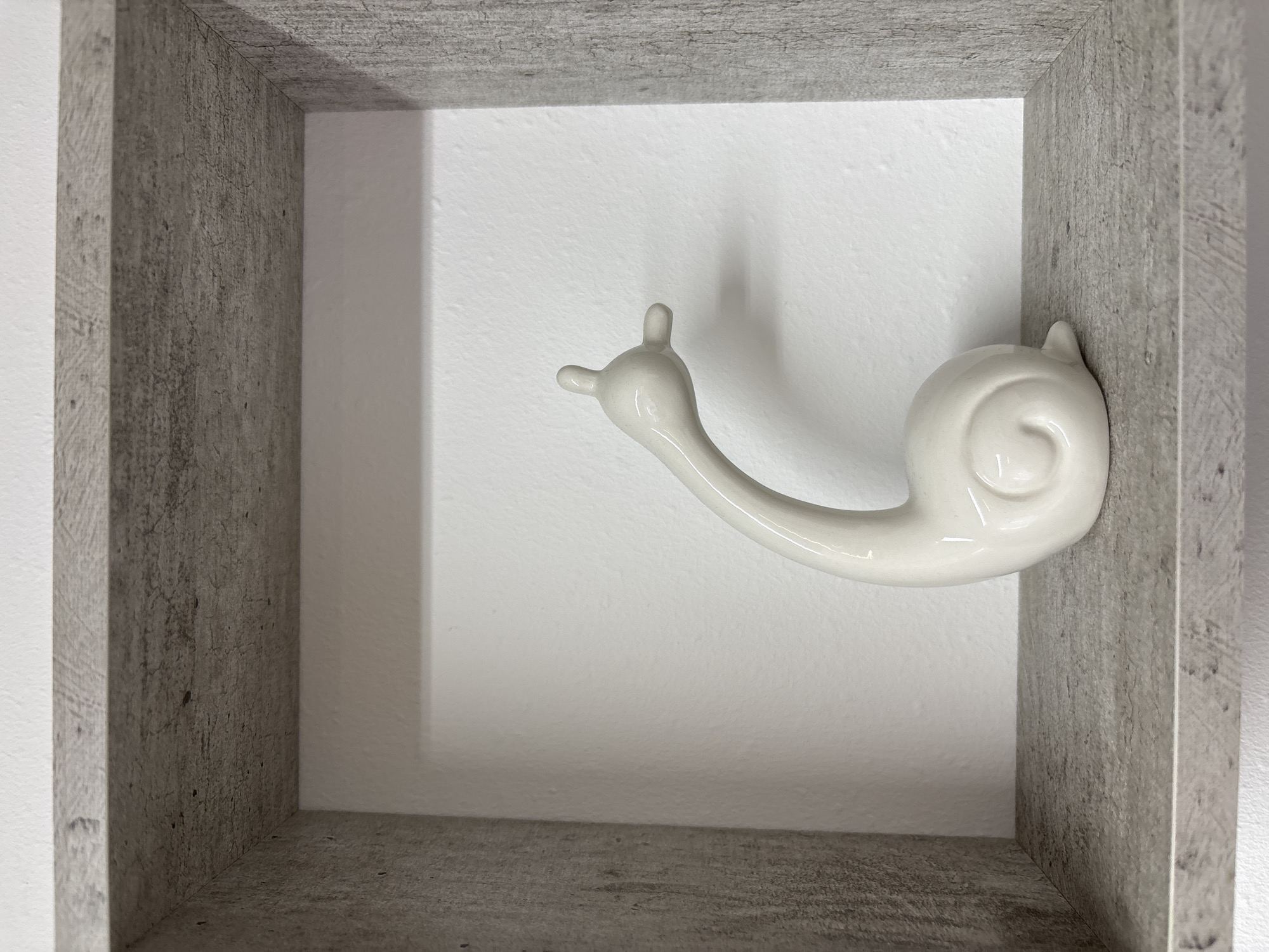 A ceramic white snail figurine is mounted on the inside wall of a grey, faux-concrete shelf. The figurine has a coiled shell and a stretched-out body with antennae. The shelf is mounted on a white wall. The image does not contain information about the location in the city of Izhevsk, Russia.