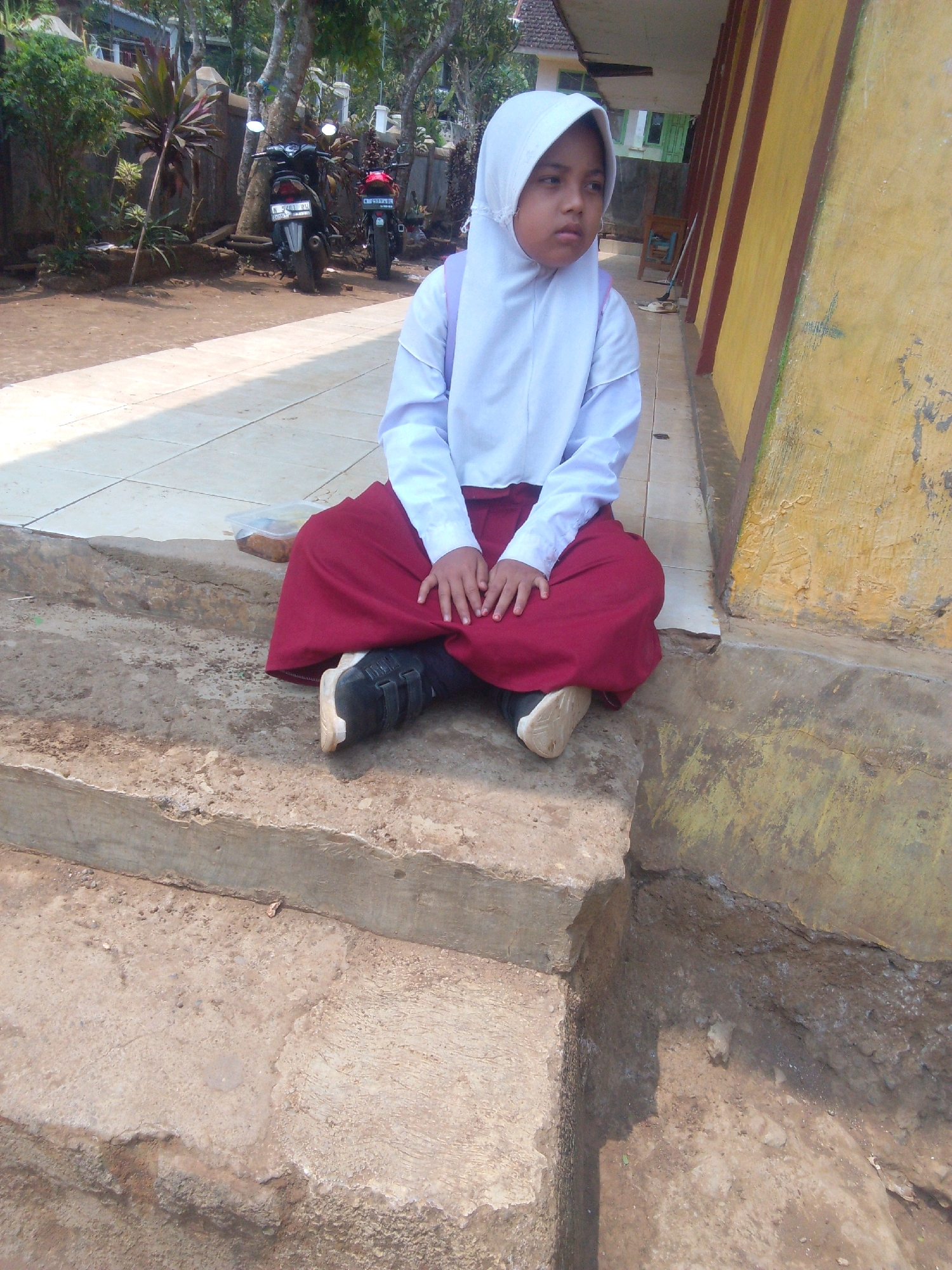 The image shows a young girl wearing a white hijab and a red skirt sitting on concrete steps outside a building. The building has a yellow wall with brown trim. In the background, there are trees, a fence, and two motorcycles parked on a dirt path. The setting appears to be outdoors, possibly in a schoolyard or residential area in Betek, Indonesia, given the presence of motorcycles and the tropical vegetation. The girl is looking to her right with a neutral expression.